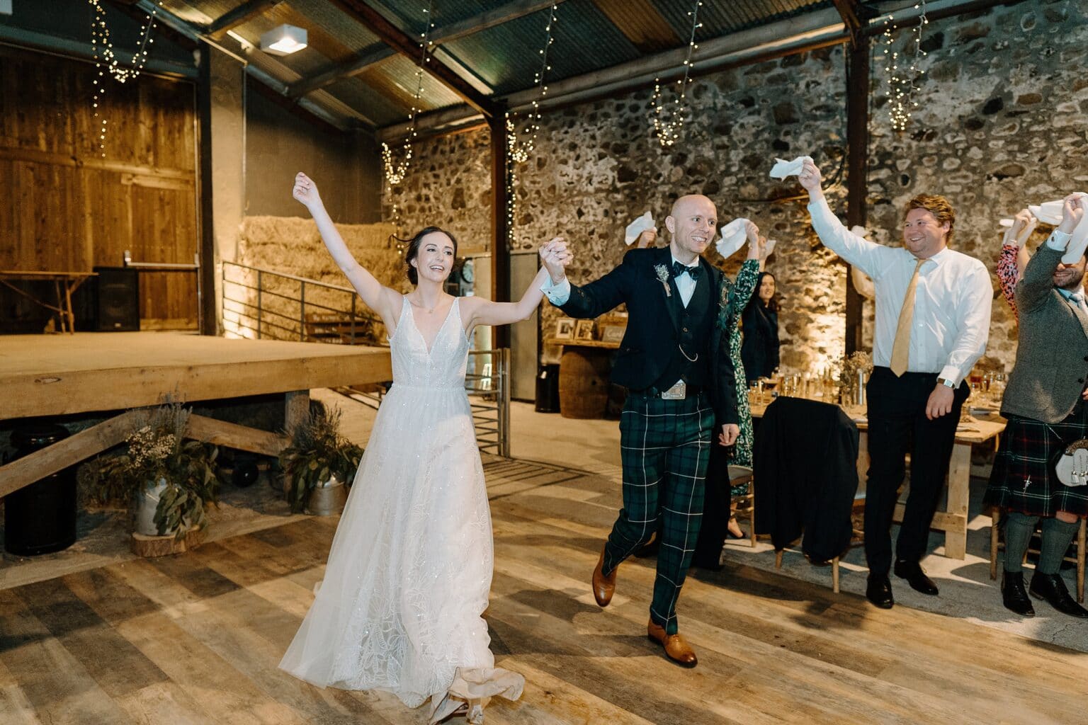 in a stone walled room with festoon lighting and hay bales in the background guests stand and wave white napkins as the bride and groom arrive for their wedding dinner at their barn wedding venue near glasgow scotland