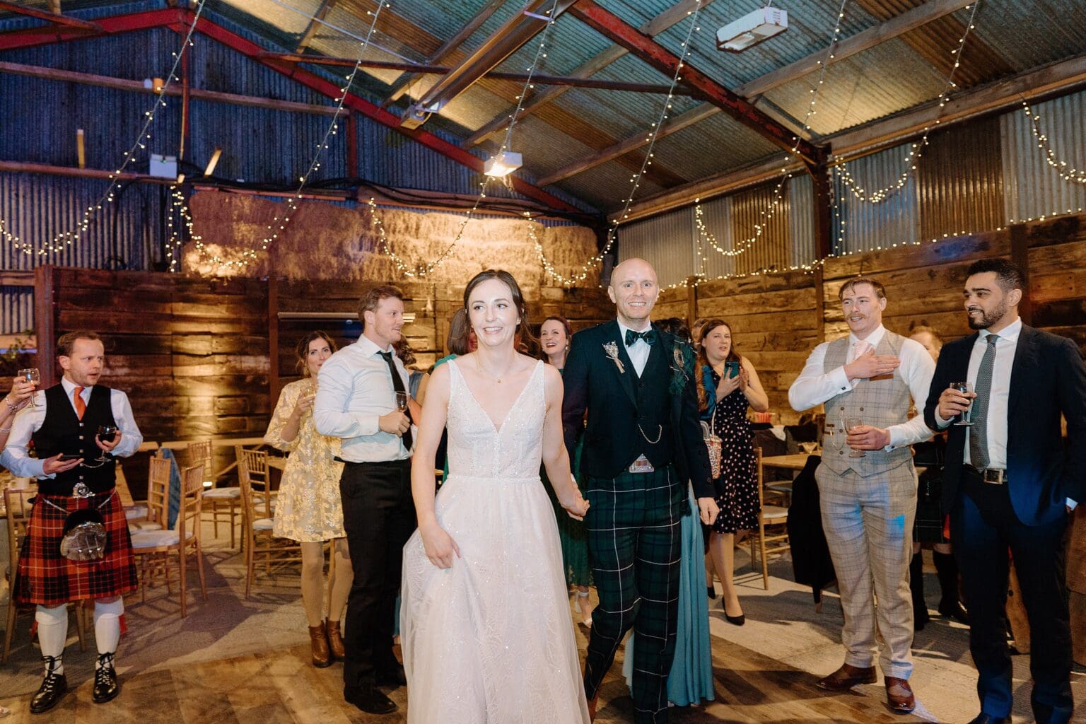 at their farm wedding venues scotland the bride and groom hold hands and walk towards the dancefloor for their first dance as guests look on beneath festoon lighting