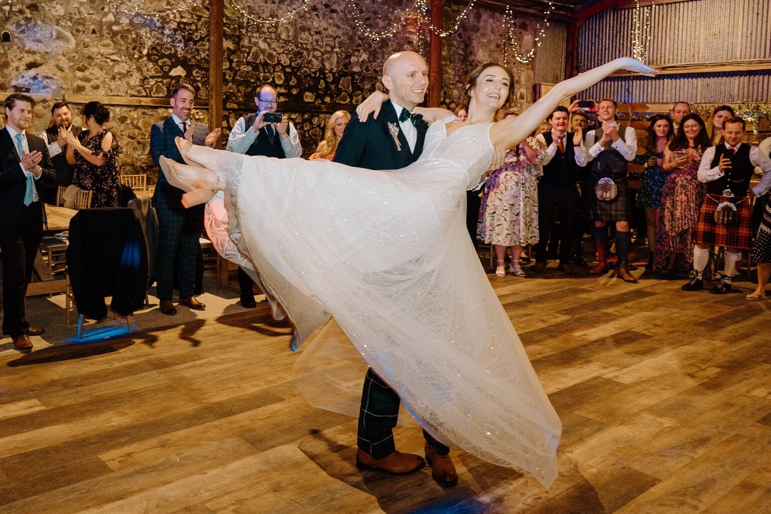 the groom lifts the bride during their first dance at their barn wedding venue as guests look on and applaud beneath festoon lighting