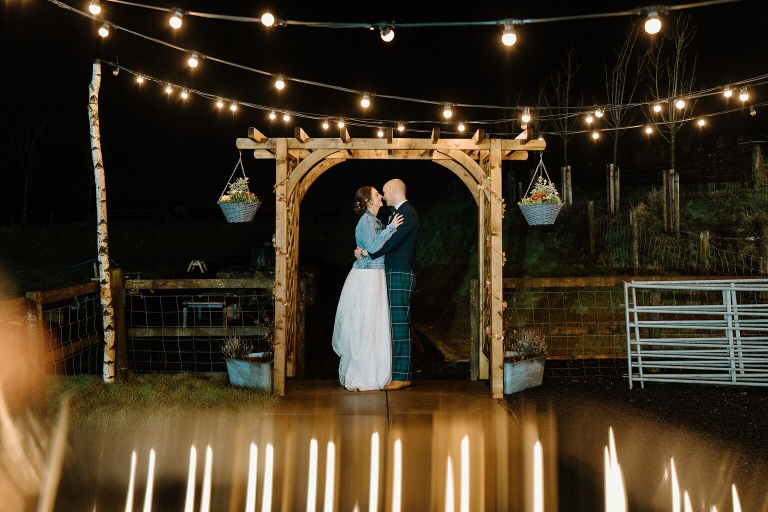 the bride and groom stand face to face beneath festoon lighting and a wooden archway with handing flower baskets at night outside their farm wedding venues scotland