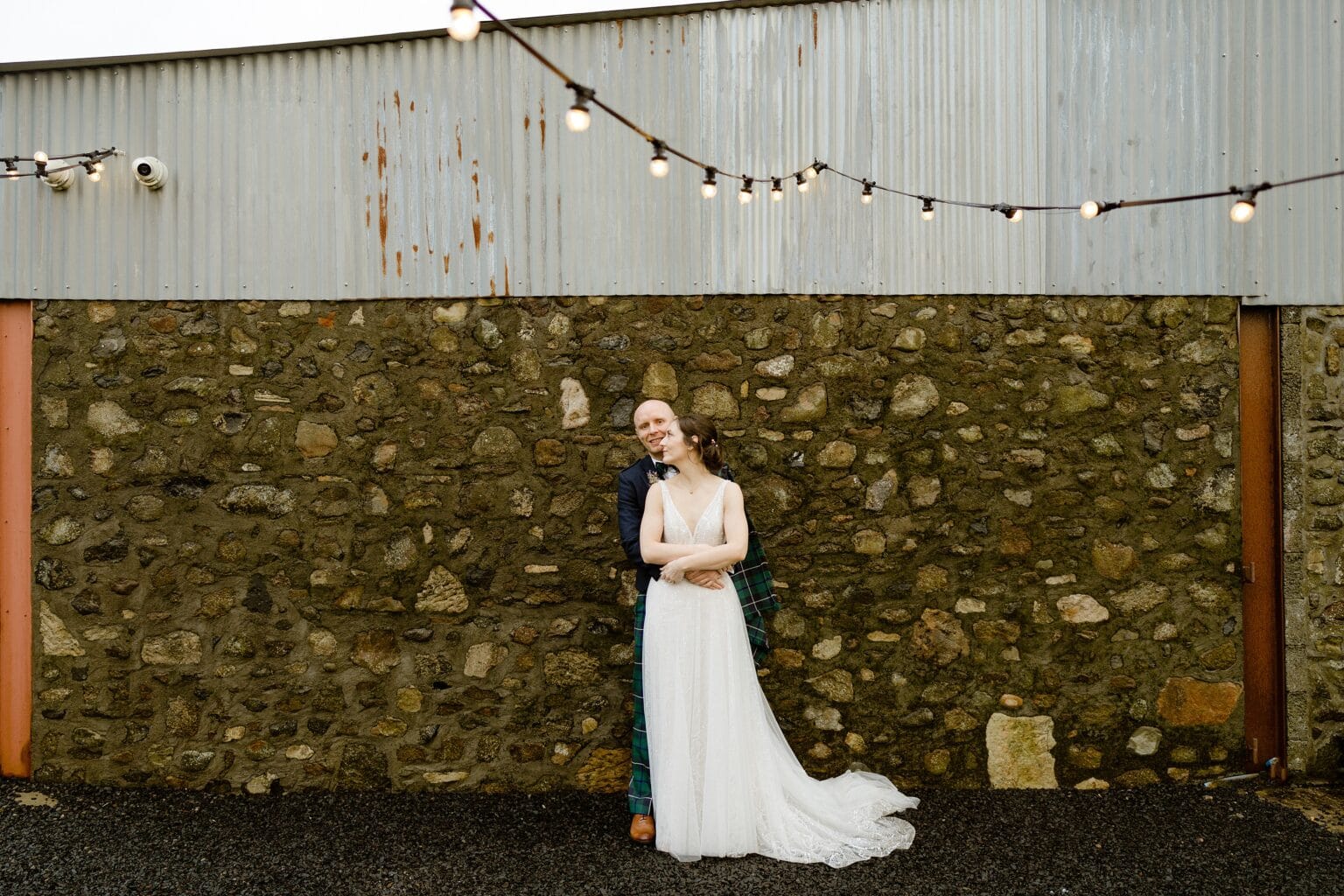 the bride and groom stand in front of a stone and corrugated iron barn wall outside their farm wedding venues scotland