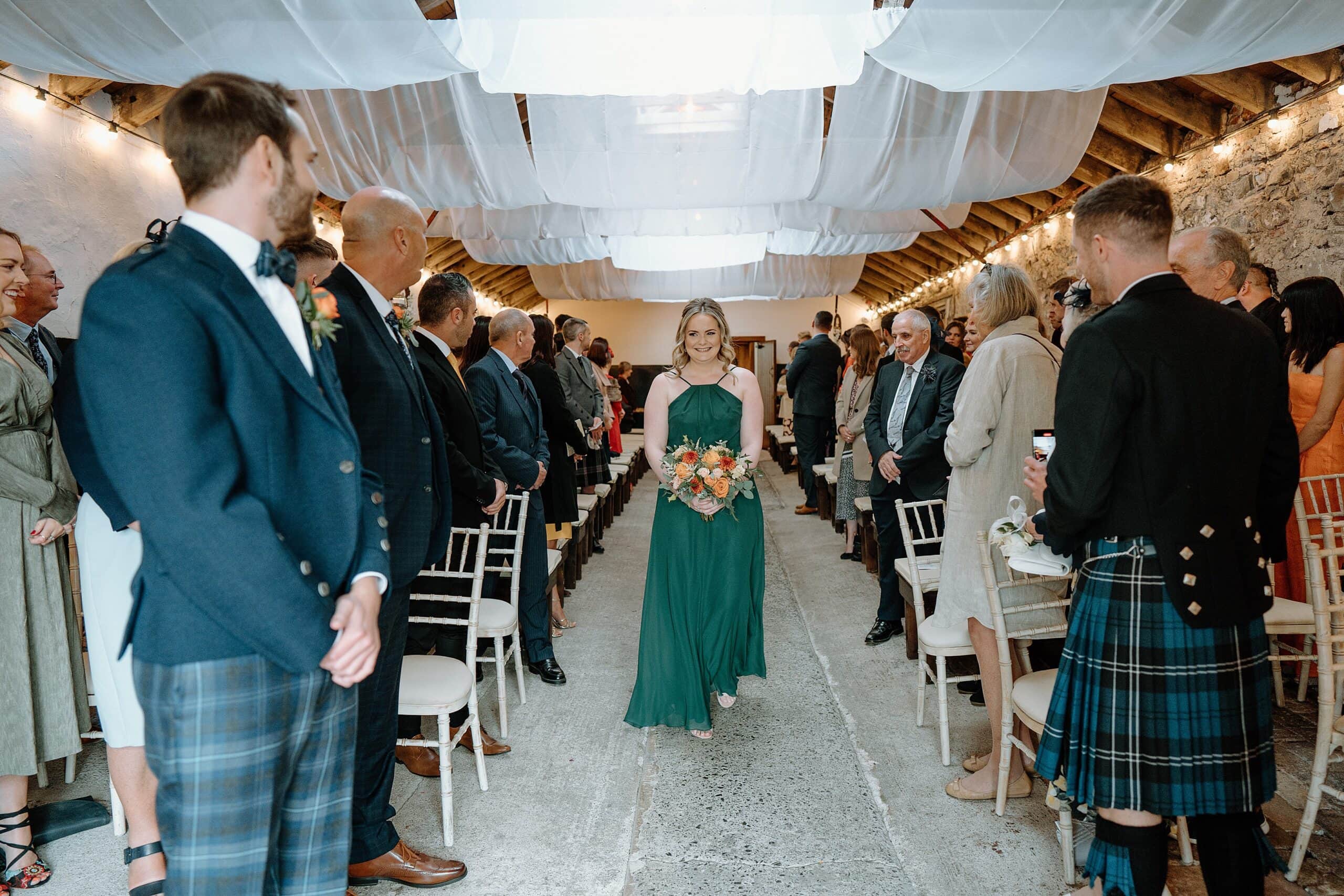 bridesmaid walks up the aisle beneath white drapes hanging from the ceiling inside the wedding byre at harelaw farm wedding barn venue fenwick scotland