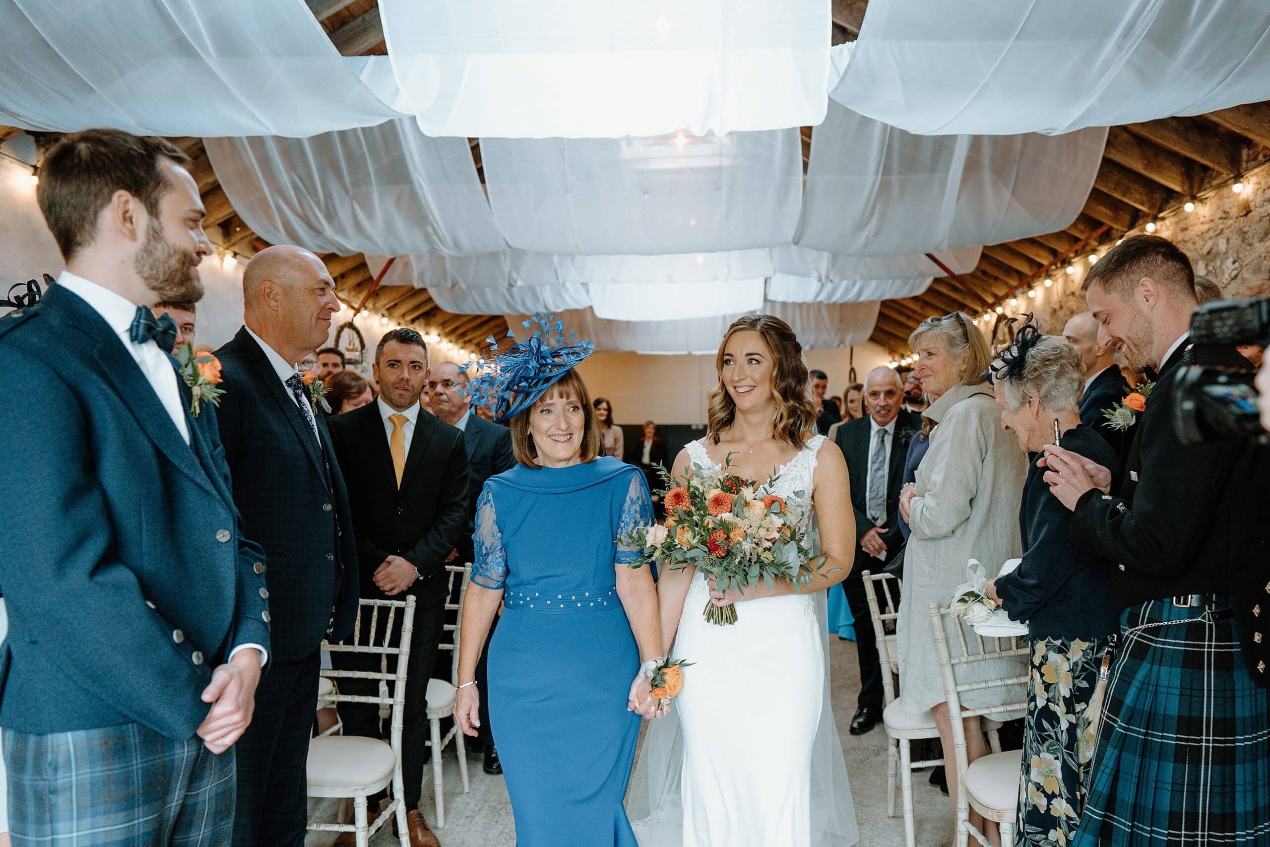 the bride and mother of the bride walk up the aisle beneath white drapes hanging from the ceiling inside the wedding byre at harelaw farm wedding barn venue fenwick scotland