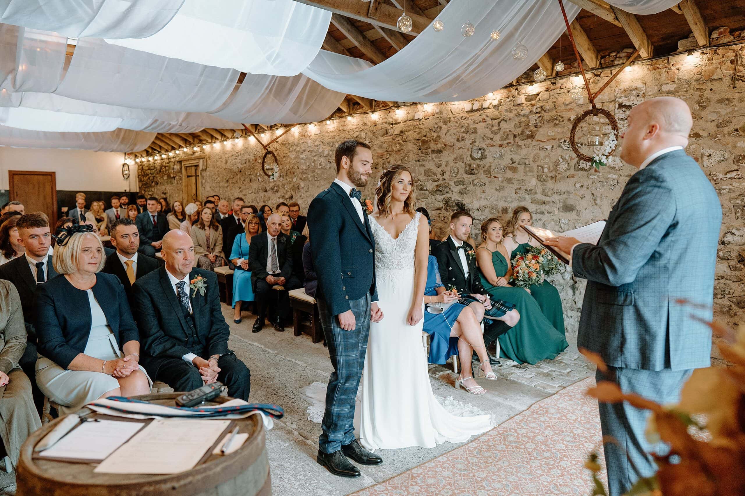 celebrant conducts wedding ceremony at harelaw farm wedding venue fenwick scotland as bride and groom listen standing beneath white ceiling drapes