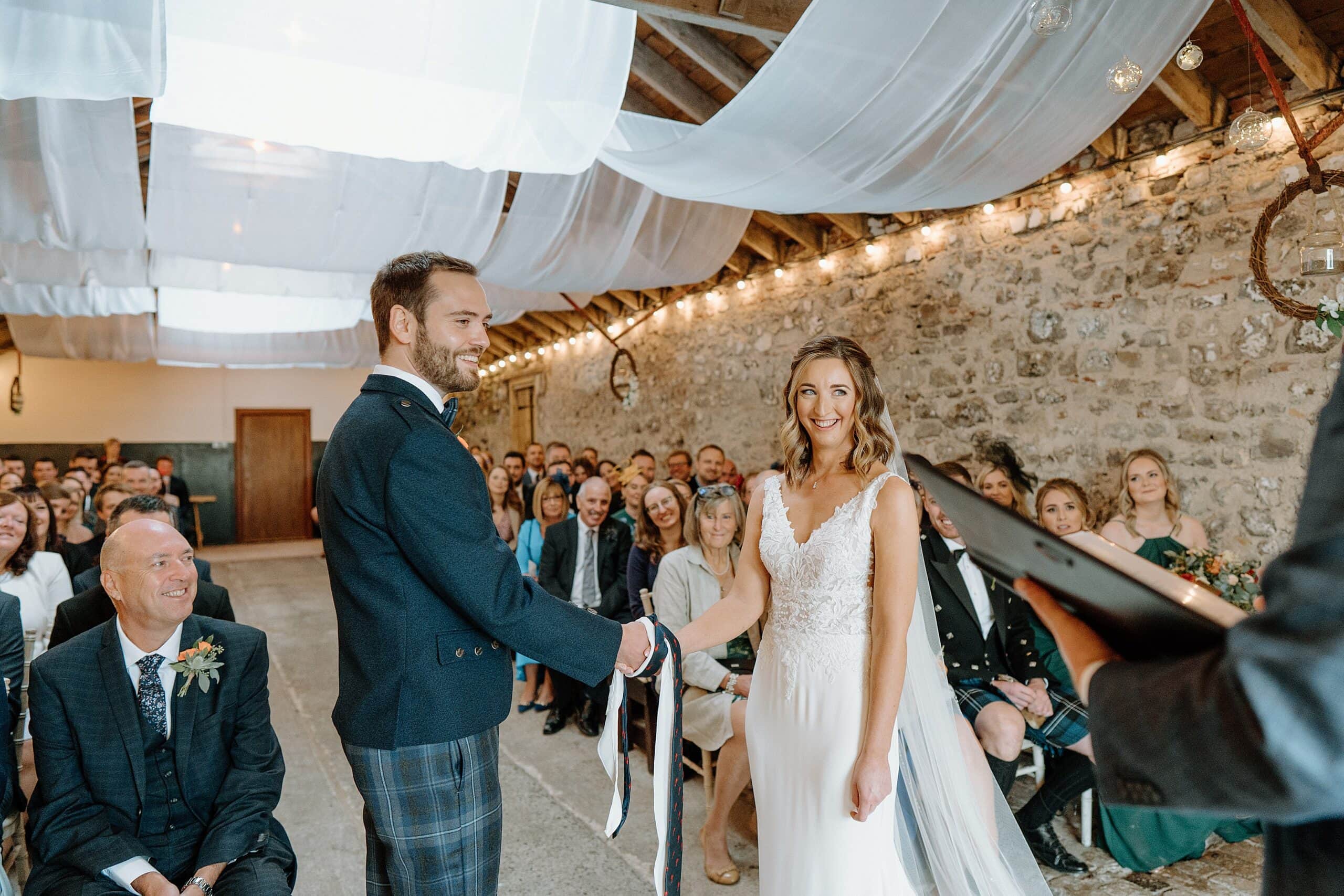 the bride and groom in handfasting ceremony beneath white drapes hanging from the ceiling during wedding ceremony at the wedding byre at harelaw farm wedding barn venue fenwick scotland