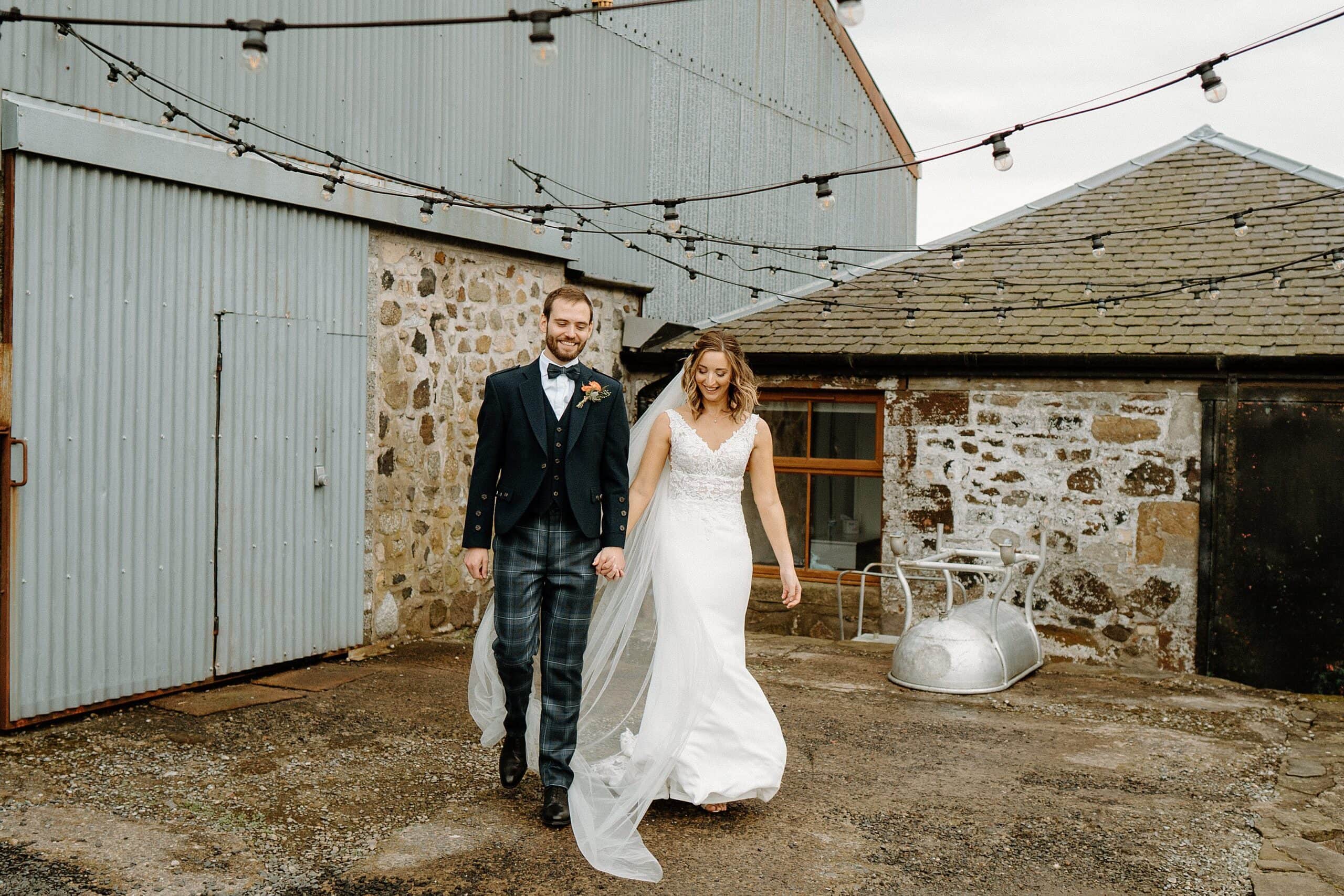 outside exterior view of harelaw farm wedding barn venue fenwick scotland showing bride and groom smiling in front of grey barn door and beneath festoon lights