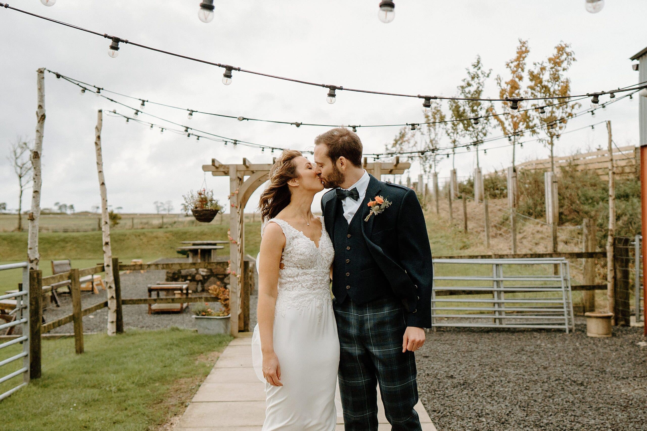 outside exterior view of harelaw farm wedding barn venue fenwick scotland showing bride and groom kissing in front of wooden arch and beneath festoon lights