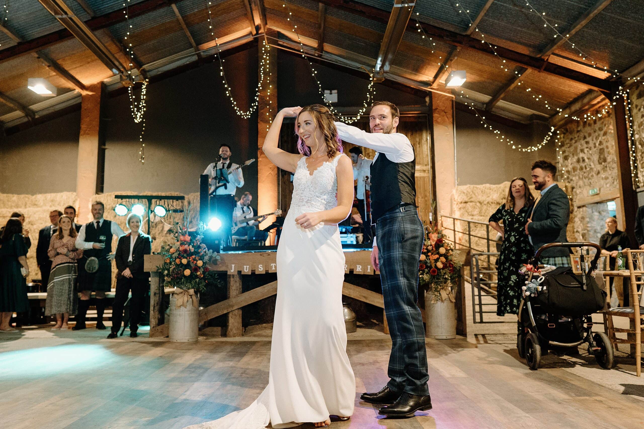 bride and groom in first dance beneath festoon lights as band play in the background in the hay barn at harelaw farm wedding venue fenwick scotland