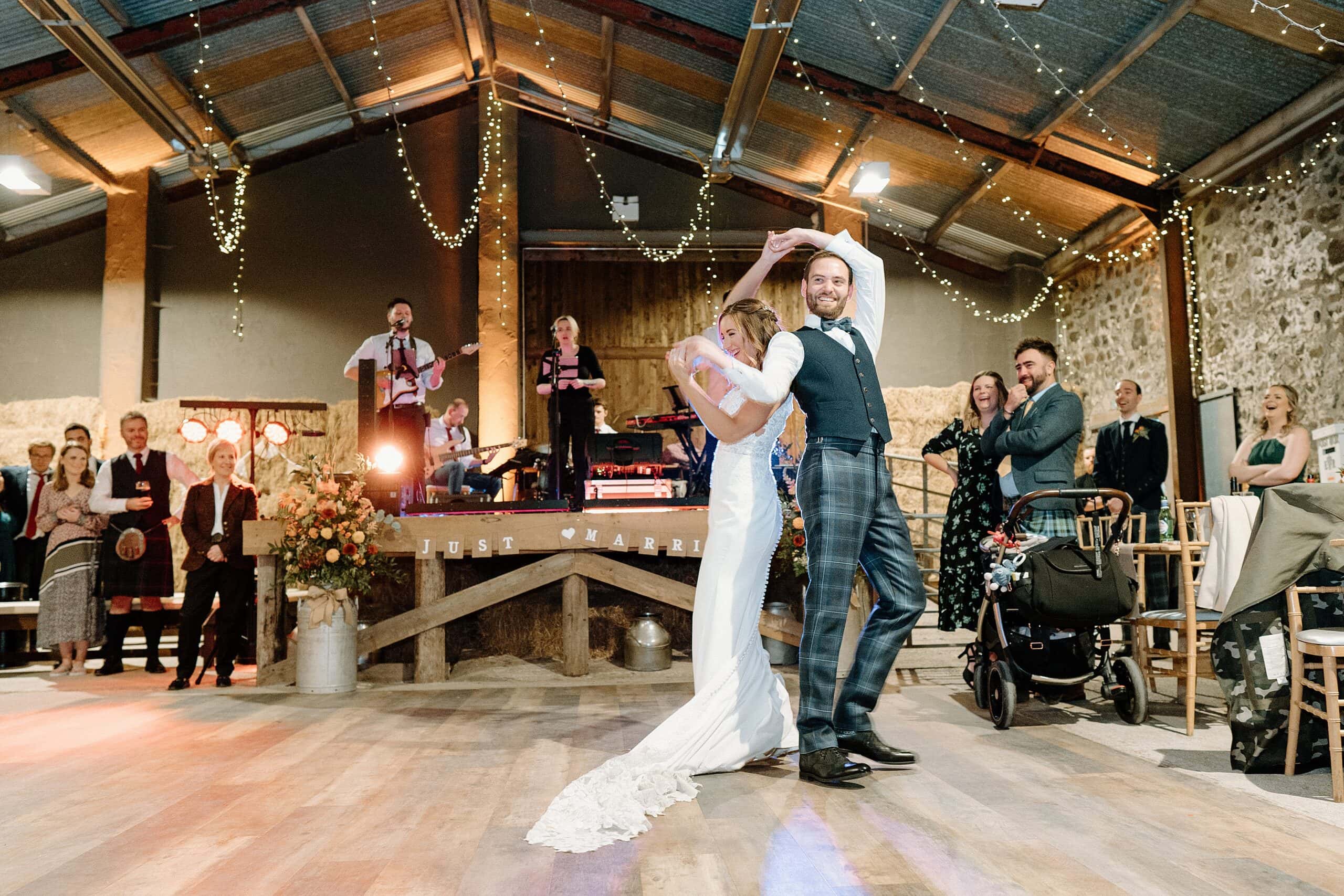bride and groom in first dance beneath festoon lights as band play in the background in the hay barn at harelaw farm wedding venue fenwick scotland
