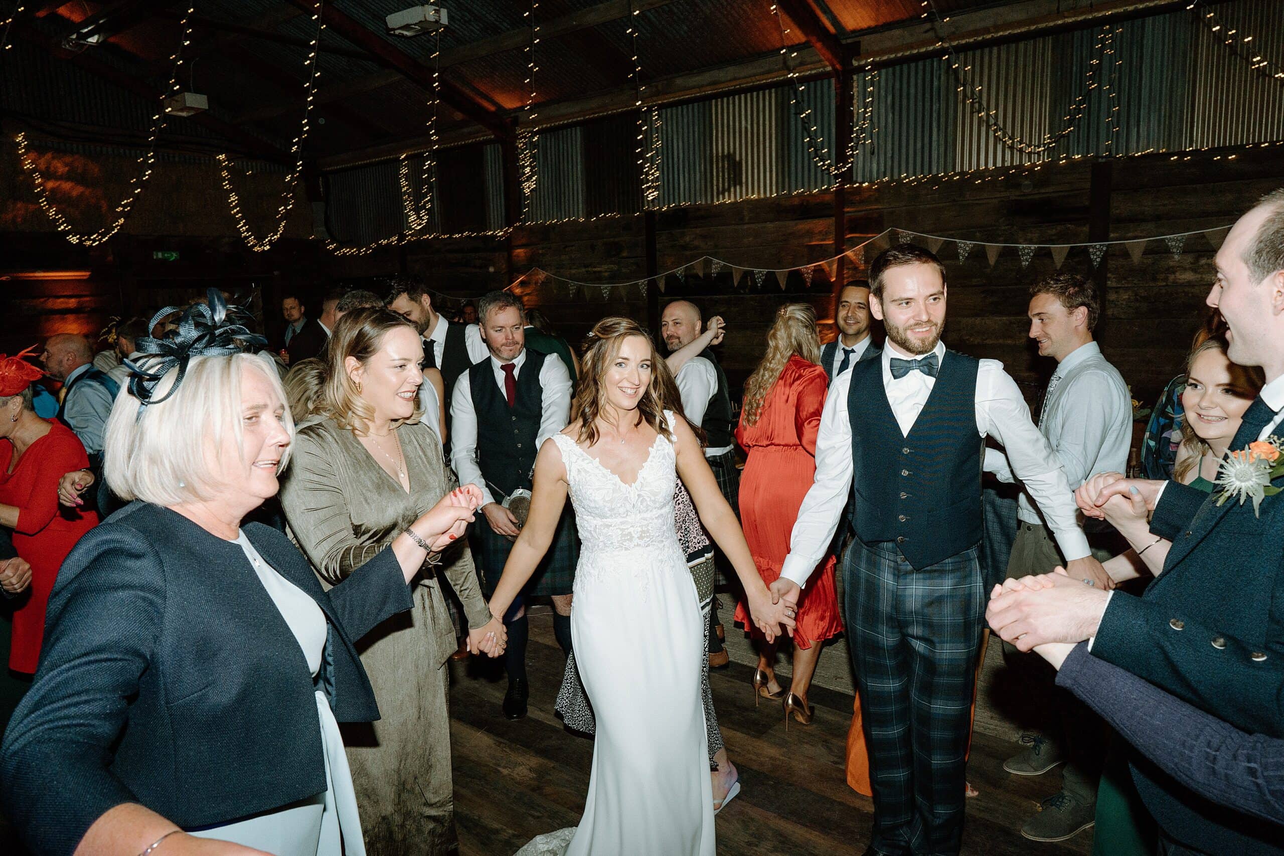 the bride and groom and wedding party dancing beneath festoon lights in the wedding barn at harelaw farm wedding venue fenwick scotland