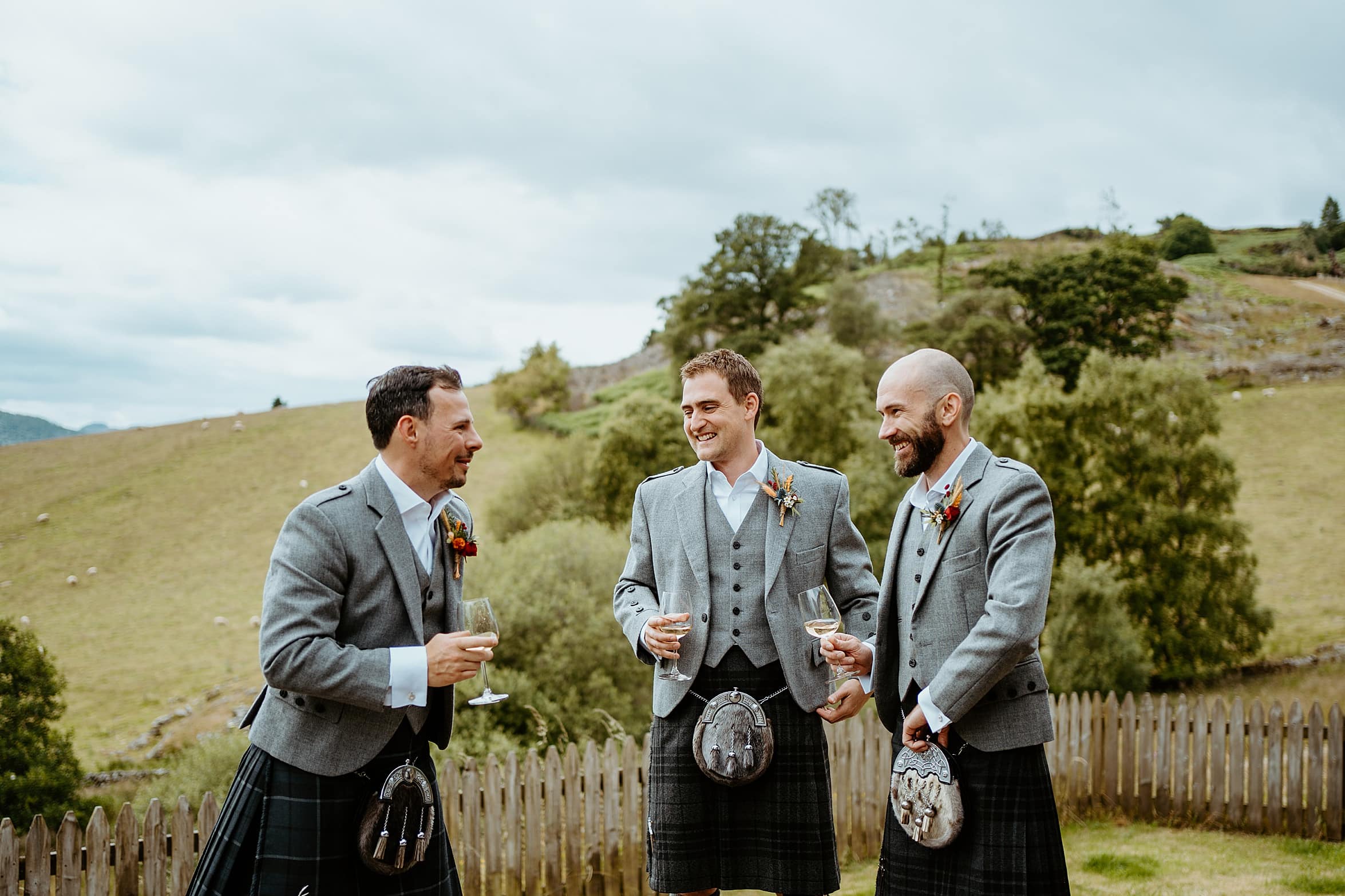 groom and groomsmen laughing as they share a drink outside with green hills and trees in background cardney steading wedding perthshire