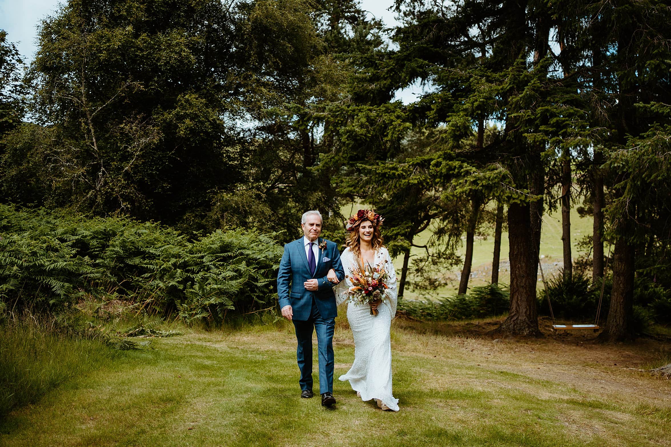 bride and father walking on grass lawn towards ceremony with woodland in background cardney steading wedding perthshire