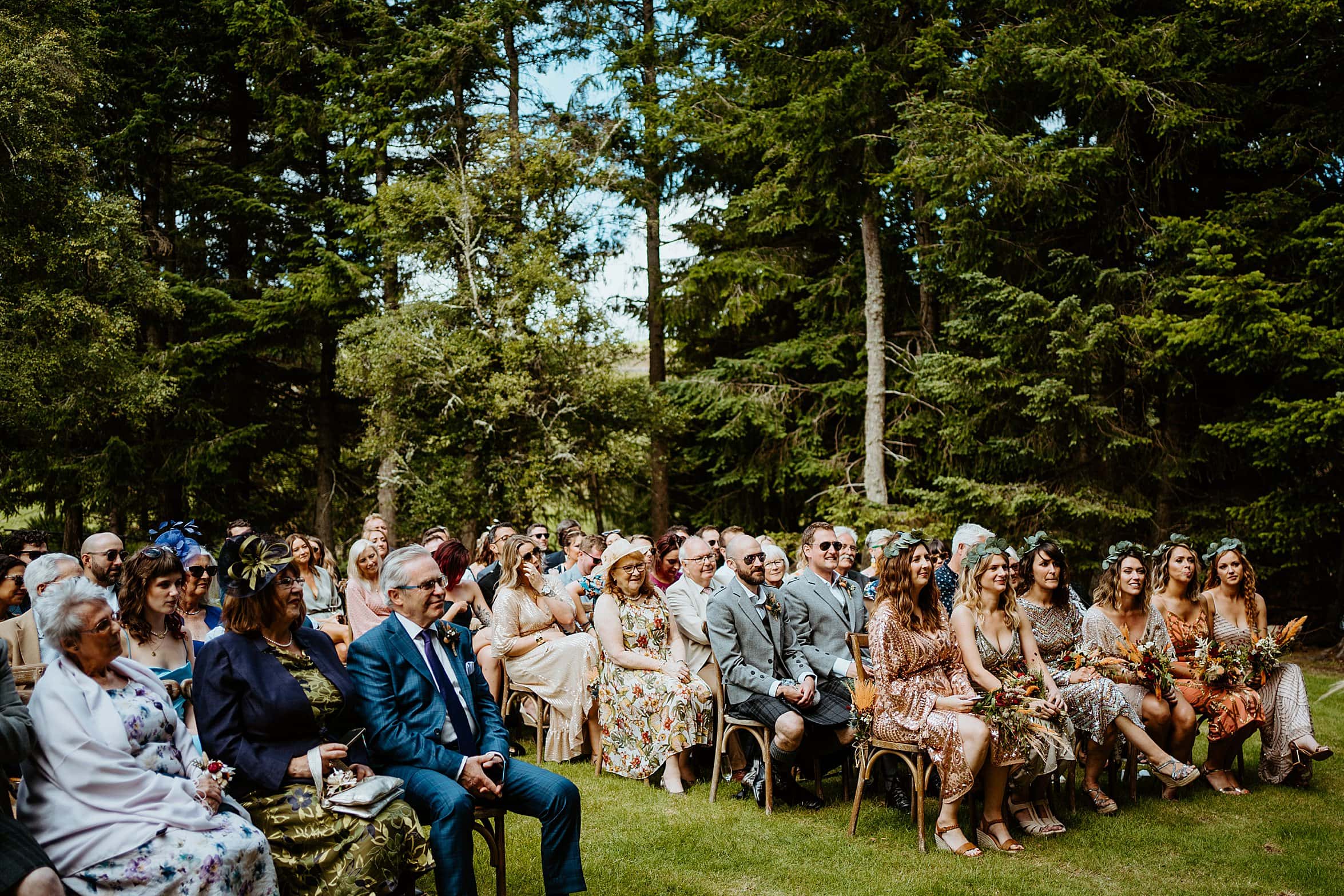 view of guests at outdoor ceremony sitting on wooden seats on grass lawn with woodland in background cardney steading exclusive use wedding venue
