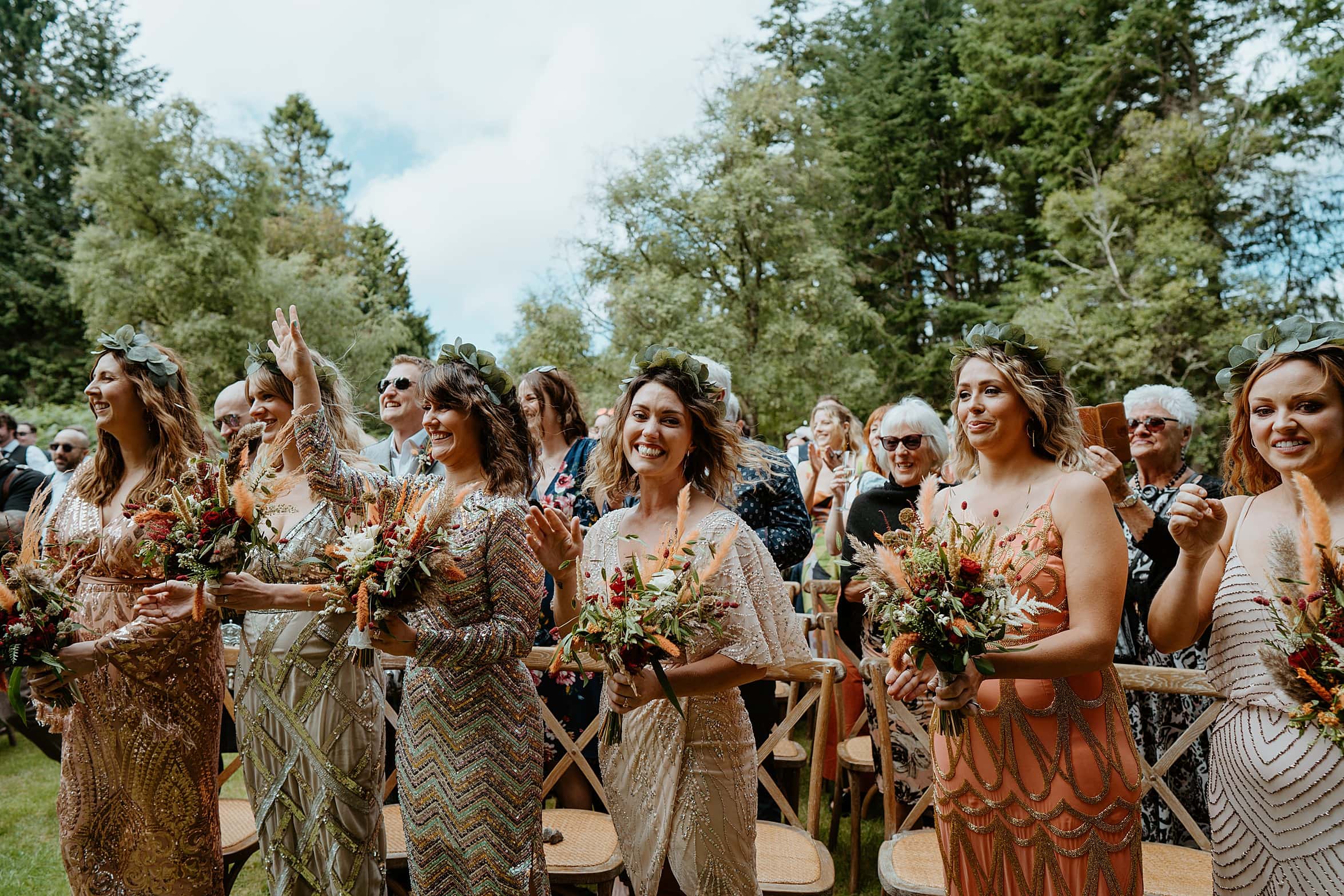 bridesmaids wearing bohemian dresses smiling at outdoor ceremony with trees and blue skies in background cardney steading wedding scotland
