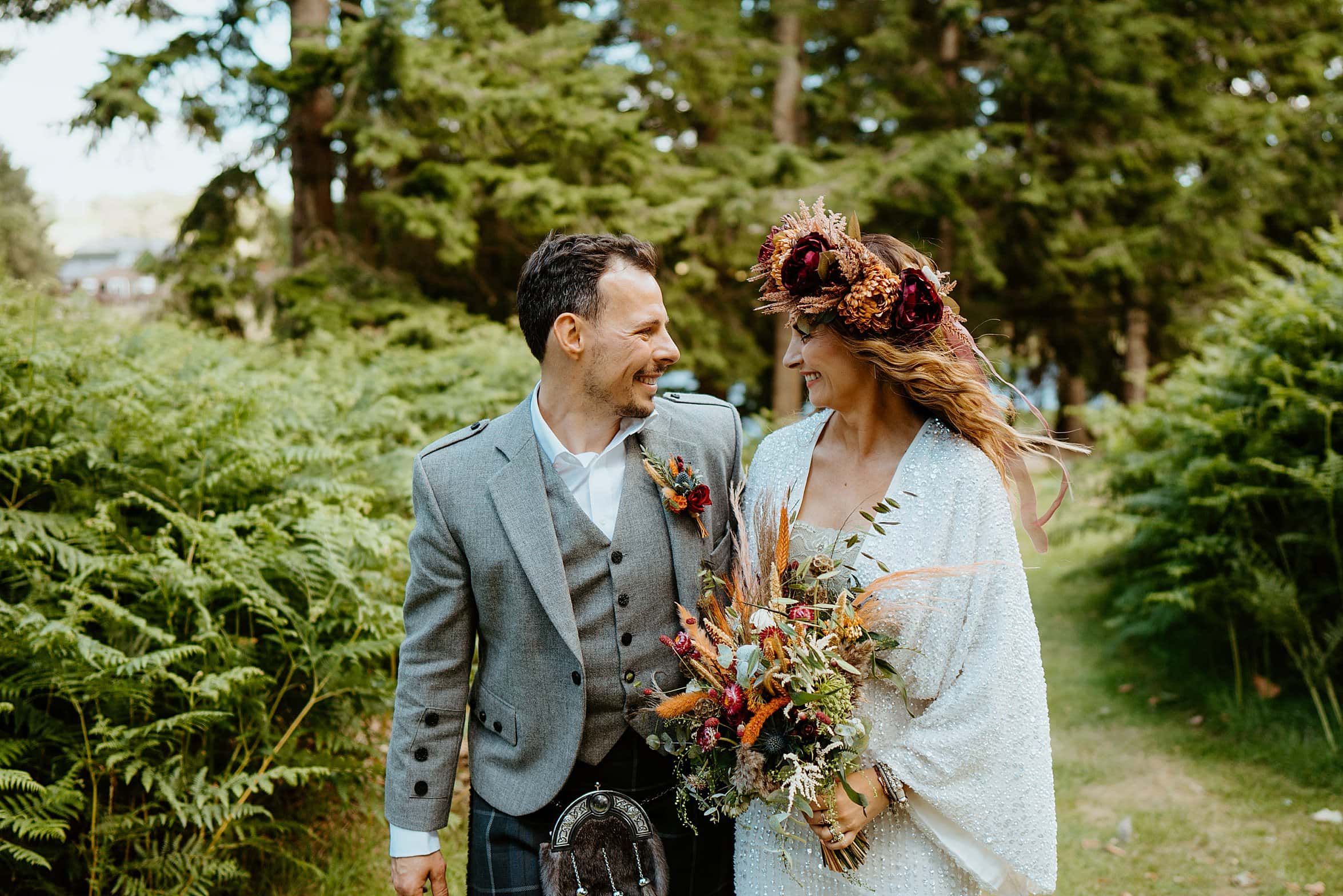bride wearing white dress floral crown and holding bouquet groom wearing kilt outfit smiling at each other as they walk through woodland cardney steading wedding venue