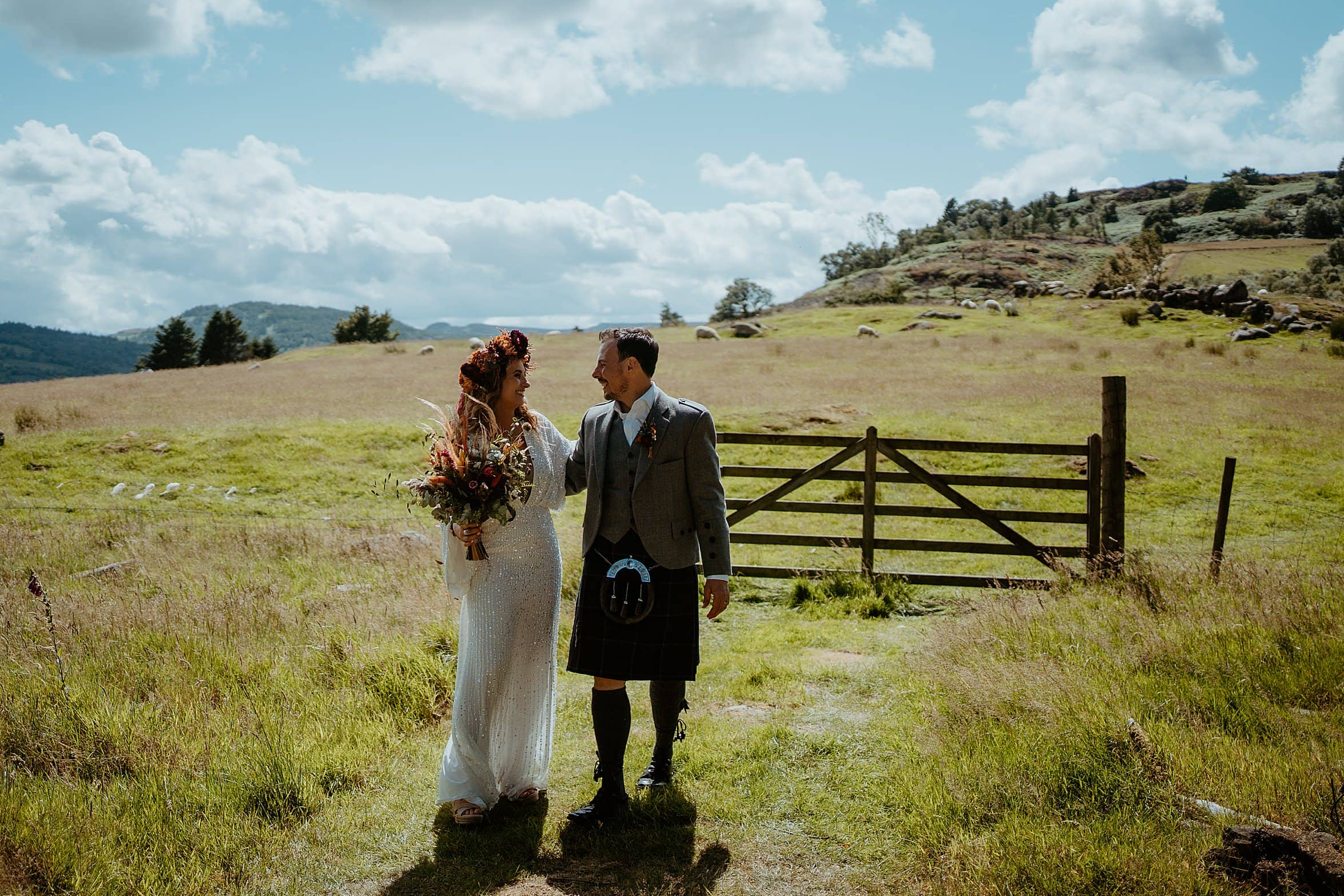 bride and groom smiling at each other as they pose in front of wooden fence in field on sunny day with blue skies cardney steading wedding perthshire