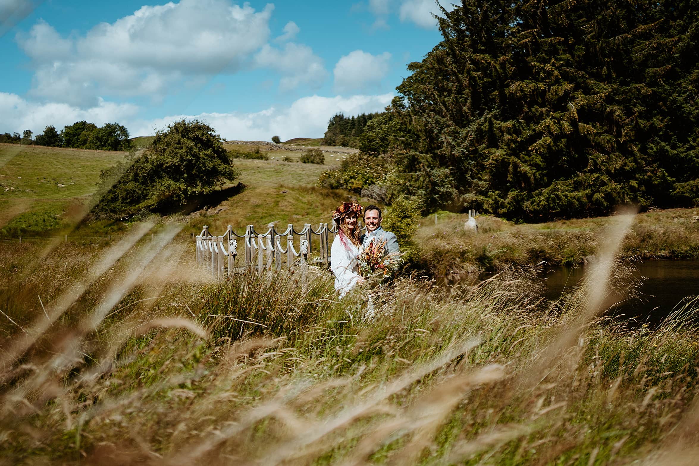 bride and groom standing on path between fields posing at camera with green trees and hills in background cardney steading exclusive use wedding venue