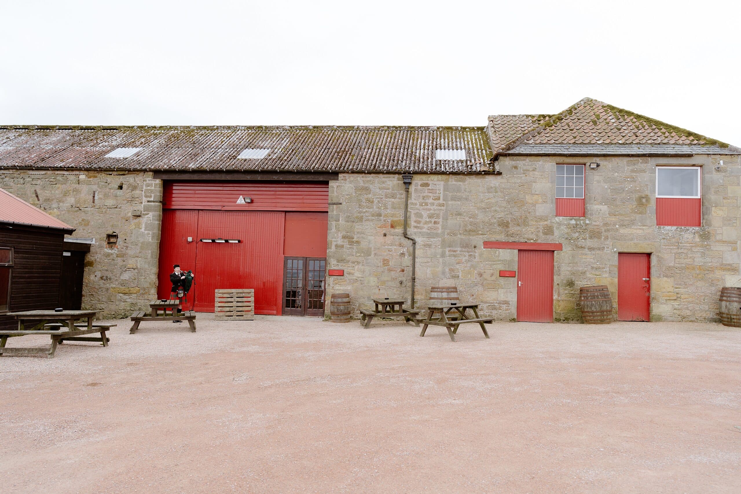 exterior outside view of kinkell byre wedding venue in st andrews scotland showing a piper with bagpipes standing in front of a red barn door