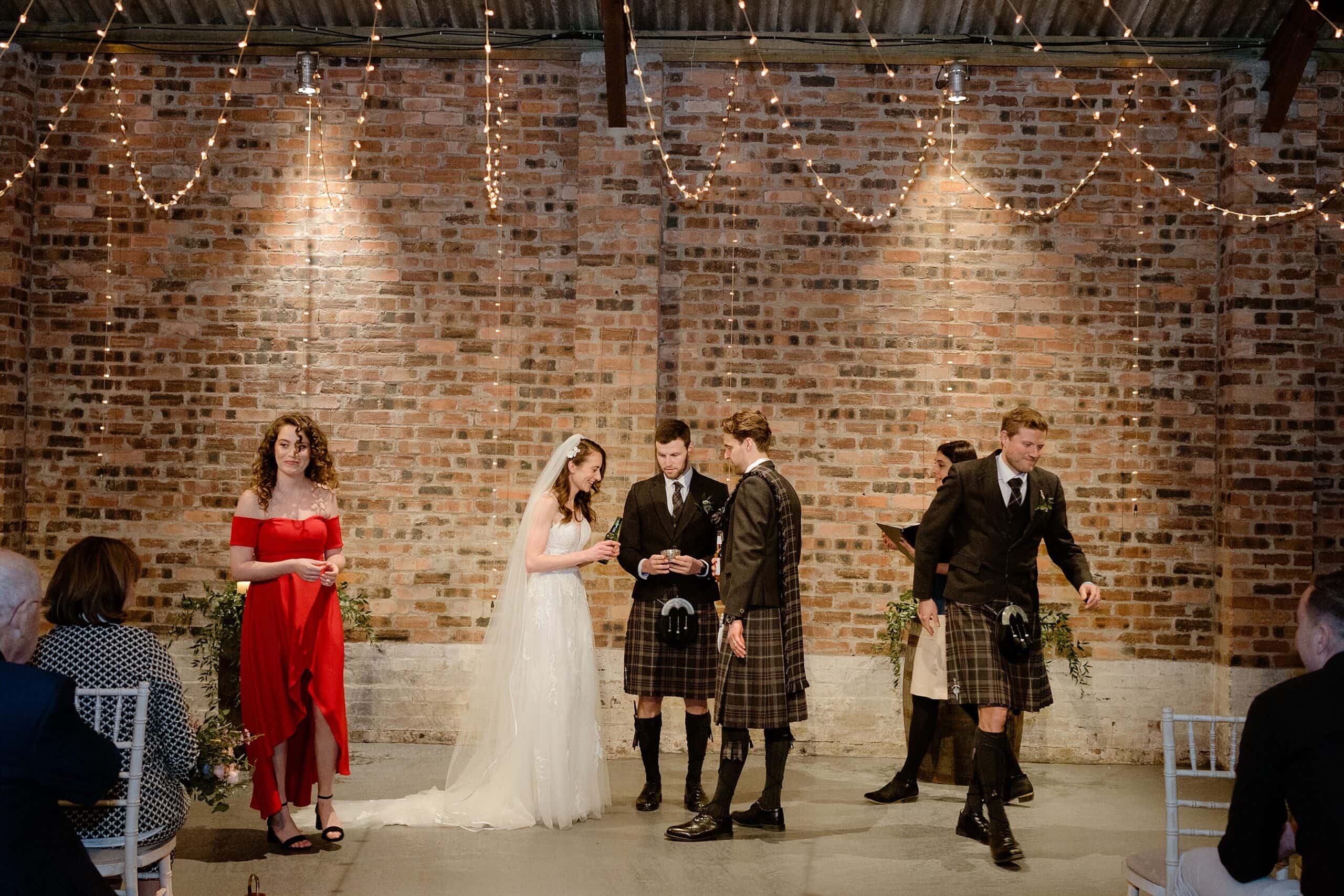 the bride and groom taking part in a quaich ceremony with a red brick wall in the background and festoon lights above in st andrews scotland