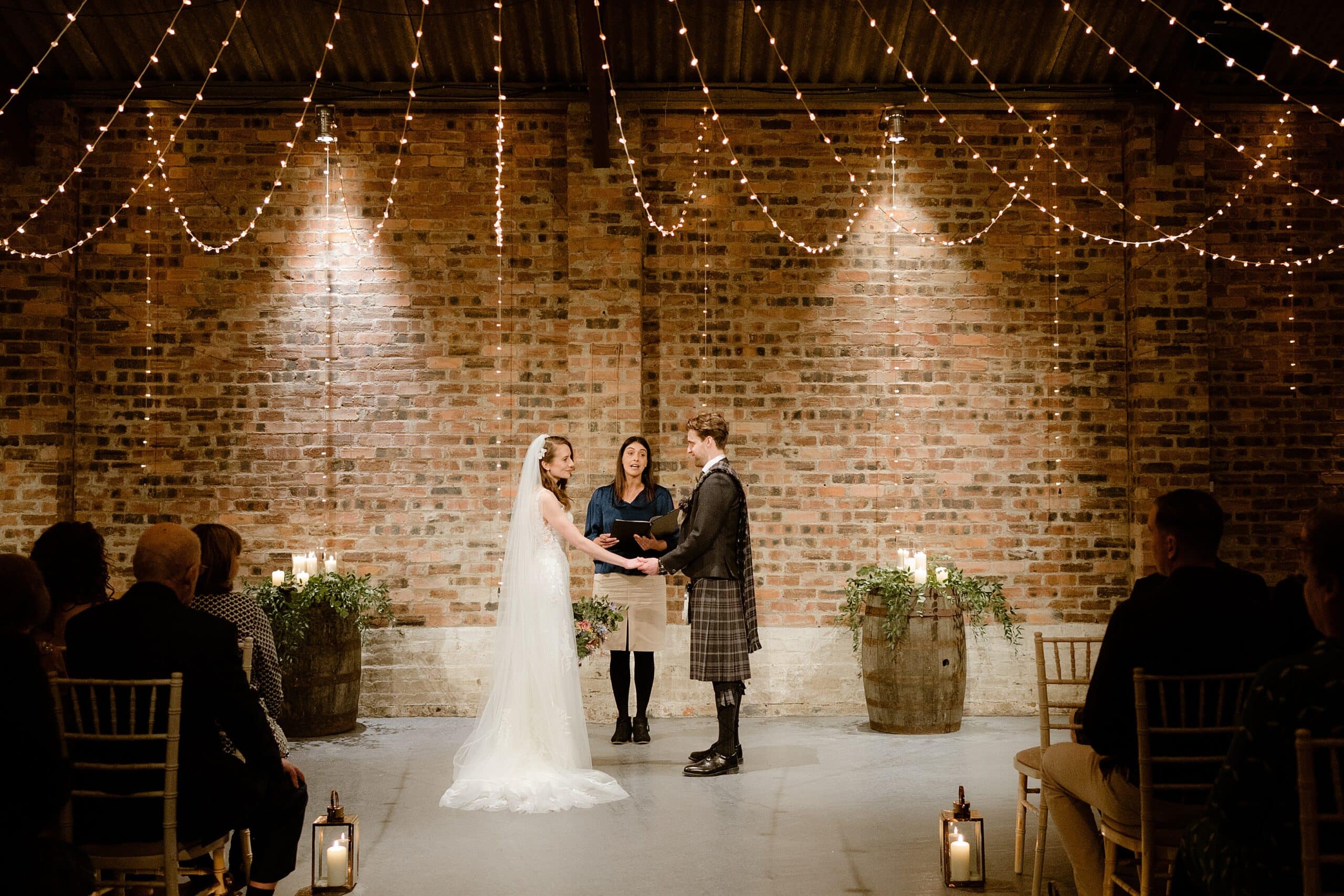 the bride and groom hold hands as celebrant conducts wedding ceremony in front of red brick wall and under festoon lights while seated guests watch at a barn wedding in st andrews scotland