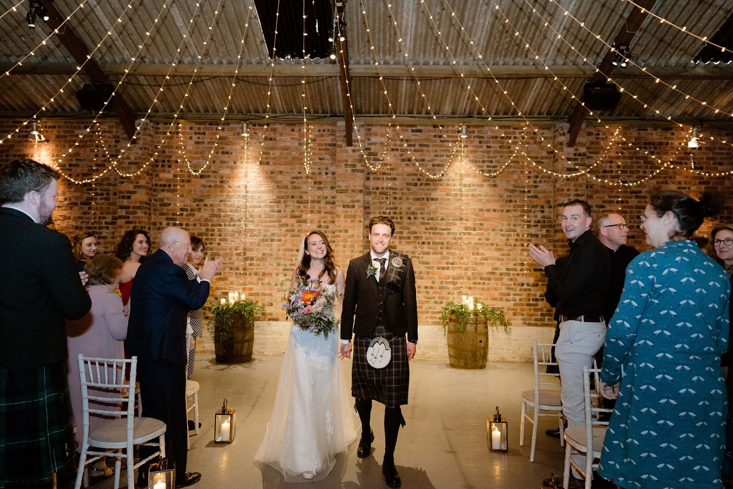 guests applaud as the bride and groom hold hands and smile as they exit down the aisle beneath festoon lights following their wedding ceremony captured by st andrews wedding photographer in scotland