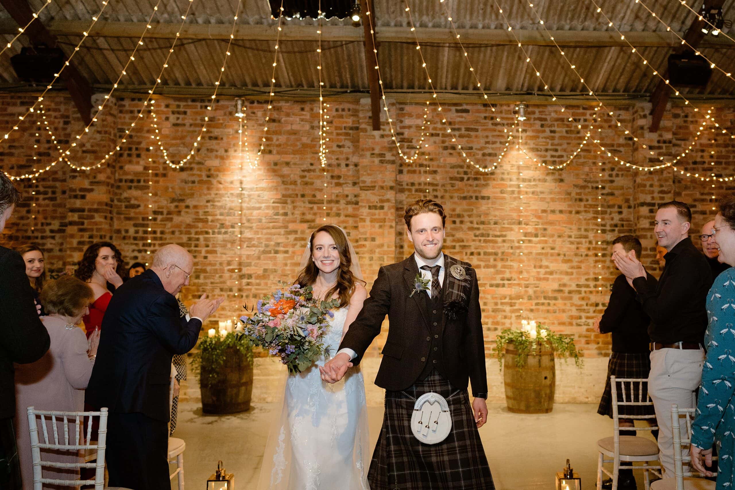 guests applaud as the bride and groom hold hands and smile as they exit down the aisle beneath festoon lights following their barn wedding ceremony captured by st andrews wedding photographer in scotland