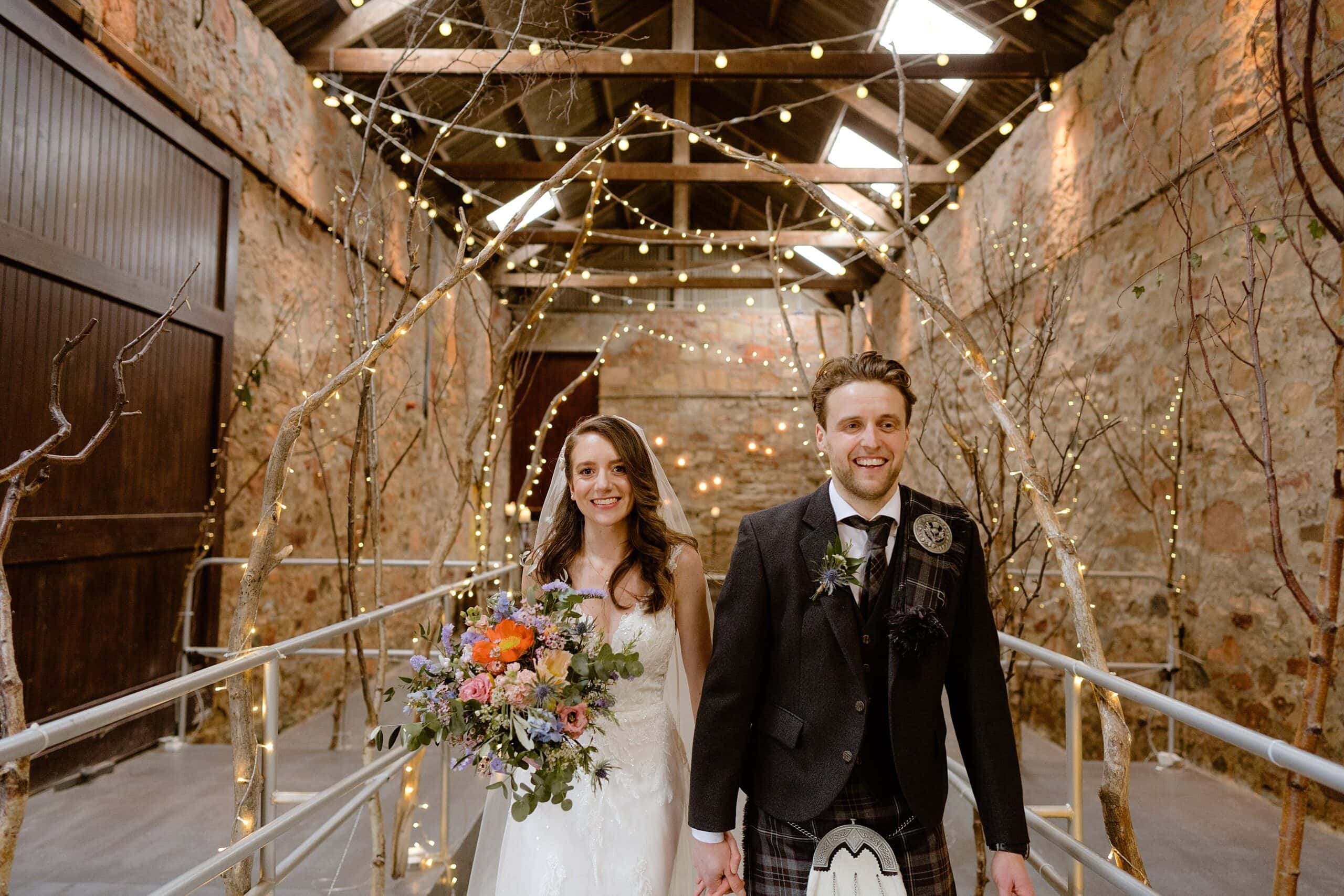 the bride and groom hold hands and smile as they walk through an archway of branches and fairy lights at a scottish barn wedding venue photographed by st andrews wedding photographer