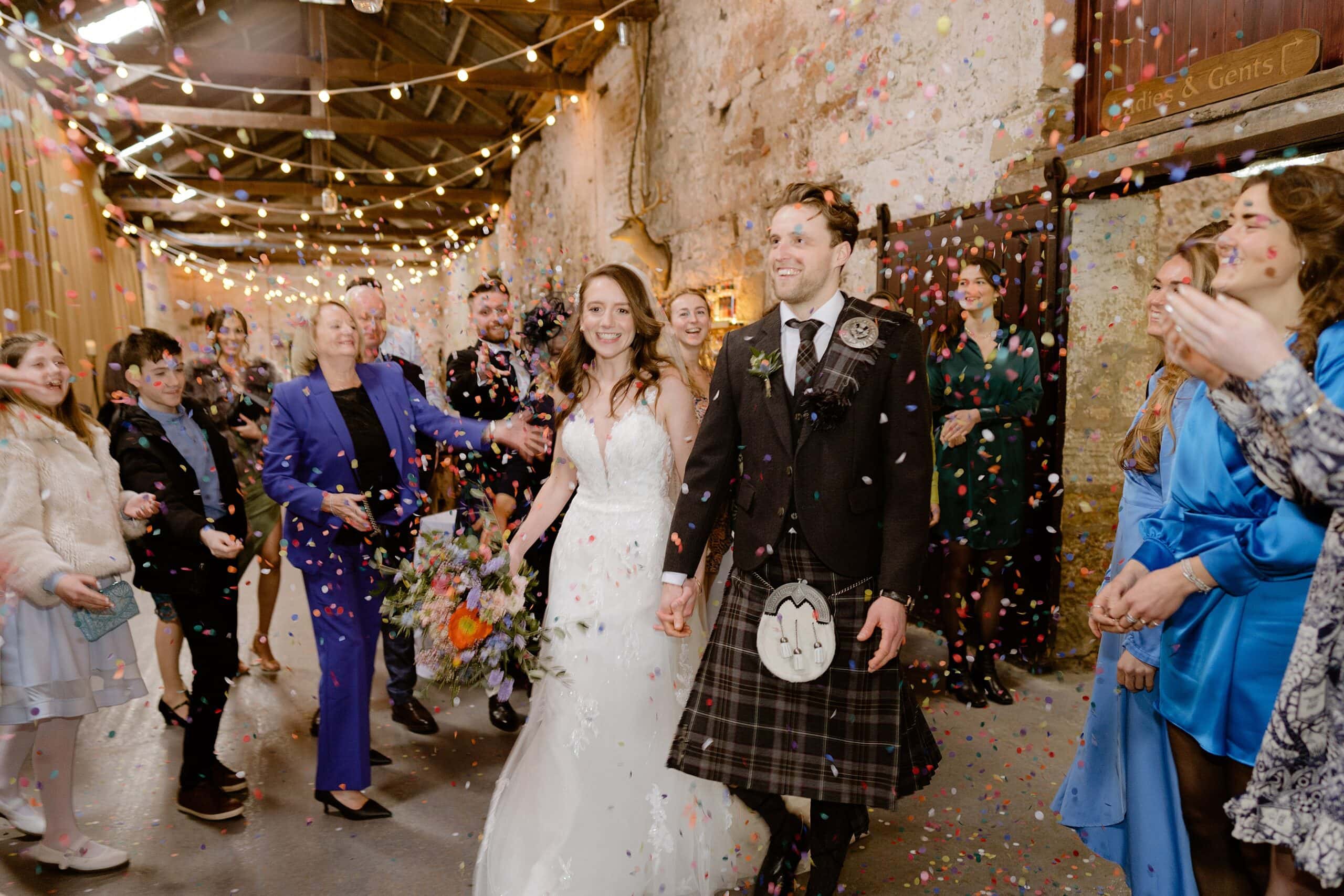 the bride and groom hold hands and smile as guests toss confetti beneath festoon lights following their wedding ceremony captured by st andrews wedding photographer