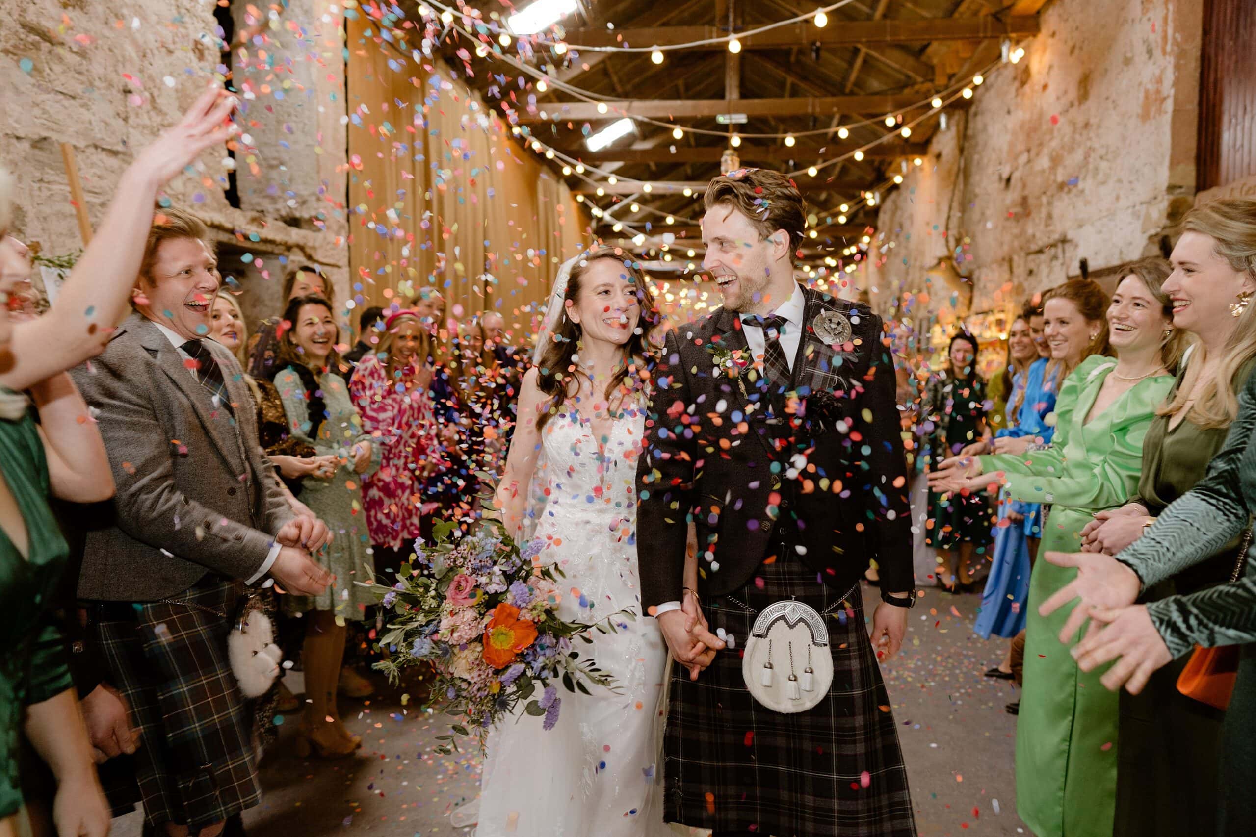 the bride and groom hold hands and smile as guests toss confetti beneath festoon lights following wedding ceremony captured by st andrews wedding photographer