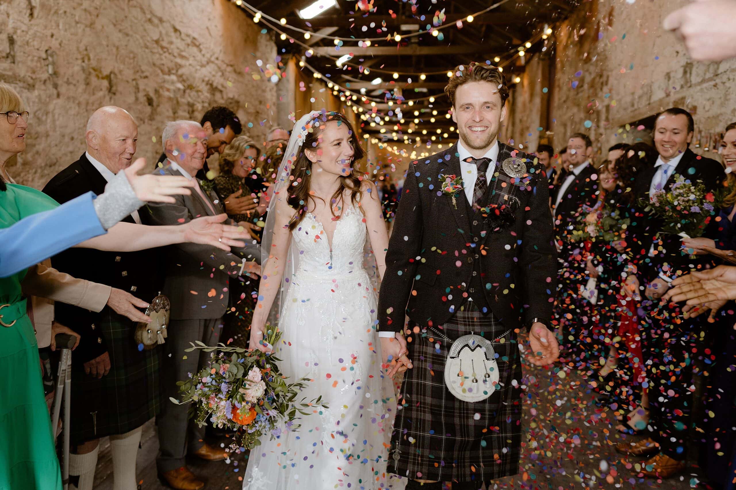 the bride and groom hold hands and smile as guests toss confetti beneath festoon lights following their wedding ceremony captured by st andrews wedding photographer
