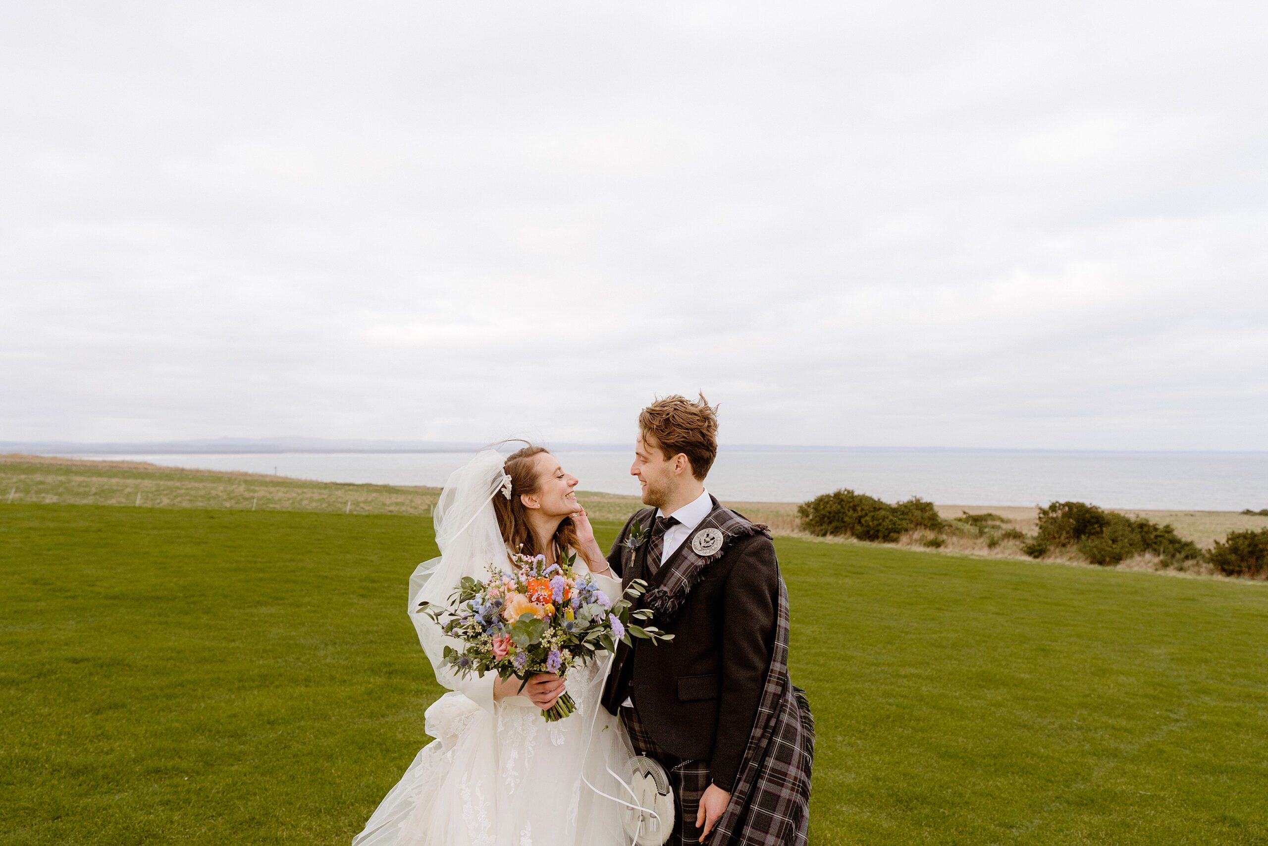 st andrews wedding photographer captures the bride and groom smiling and looking into each others eyes outside their barn wedding venue in scotland with the sea in the background