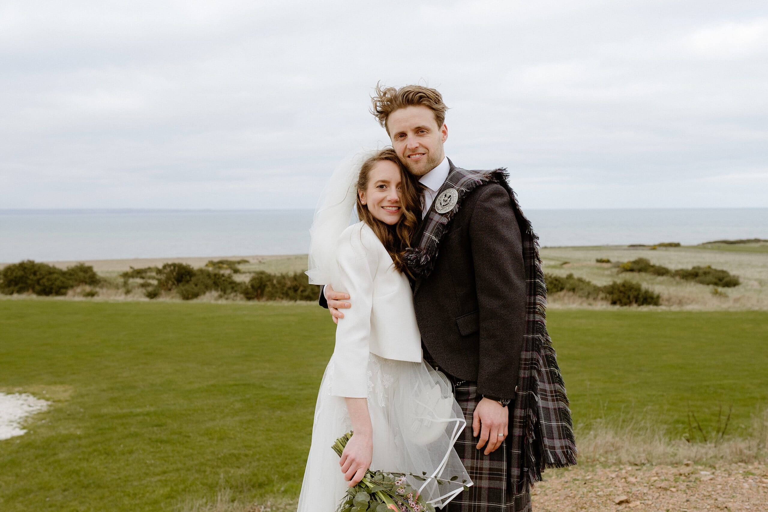 st andrews wedding photographer captures the groom with his arm around the bride outside their barn wedding venue in scotland with the sea in the background