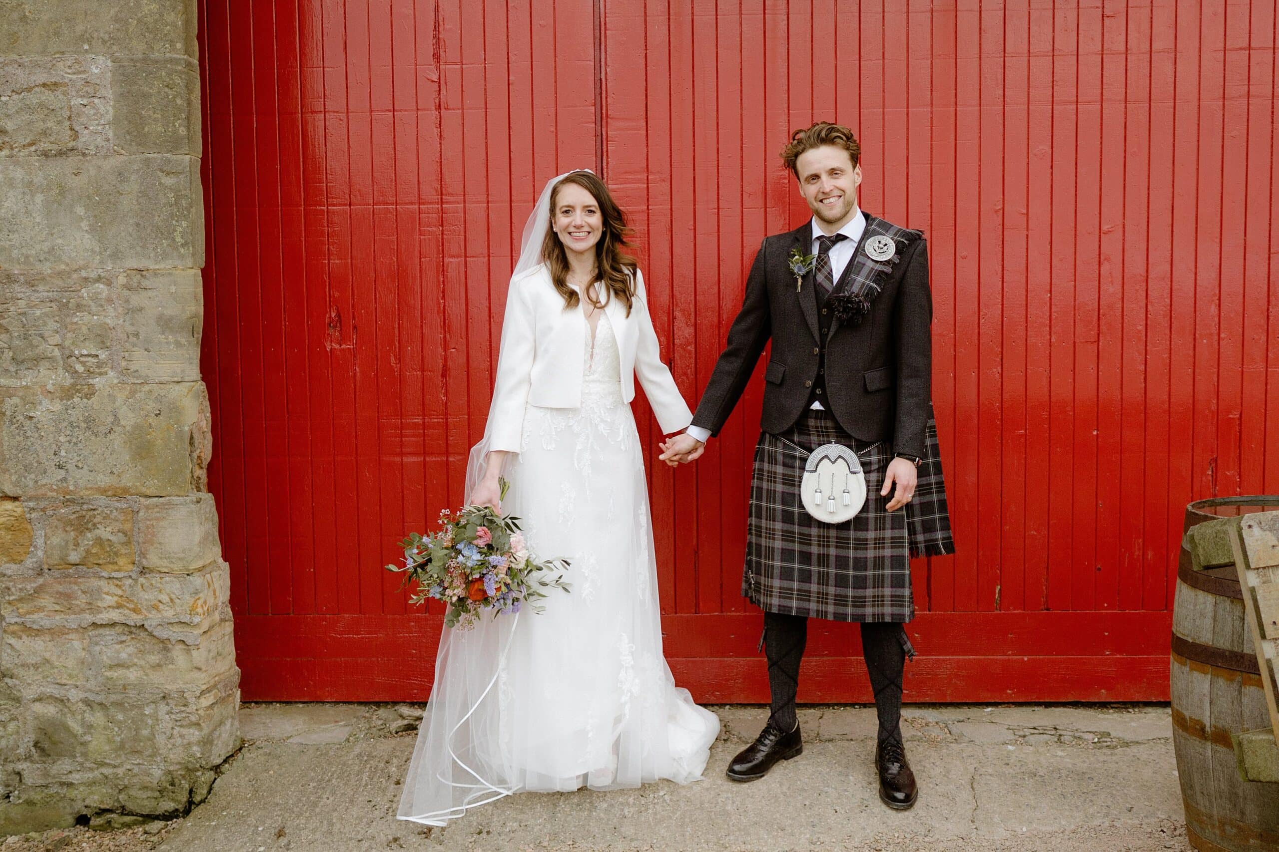 the bride and groom smile and hold hands in front of a red barn door outside kinkell byre scottish wedding venue