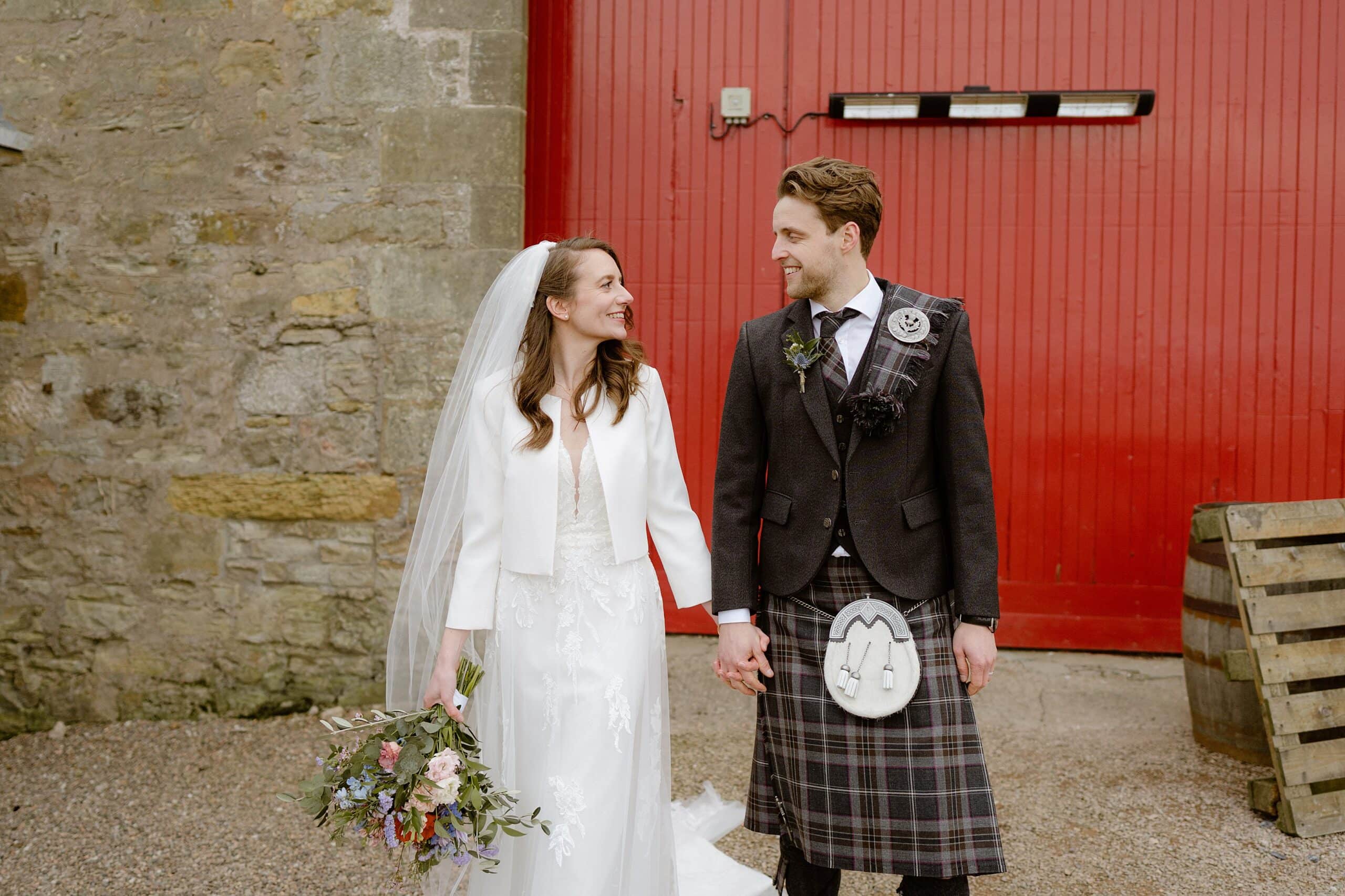 the bride and groom smile and hold hands in front of a red barn door outside their unique barn wedding venue in scotland captured by st andrews wedding photographer