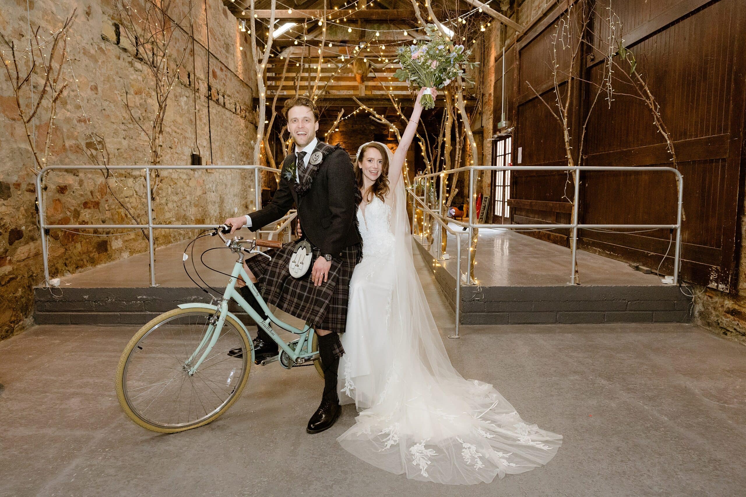 interior inside view of a barn wedding venue showing the bride and groom posing on a bicycle with an archway of branches and festoon lights in the background photographed by st andrews wedding photographer