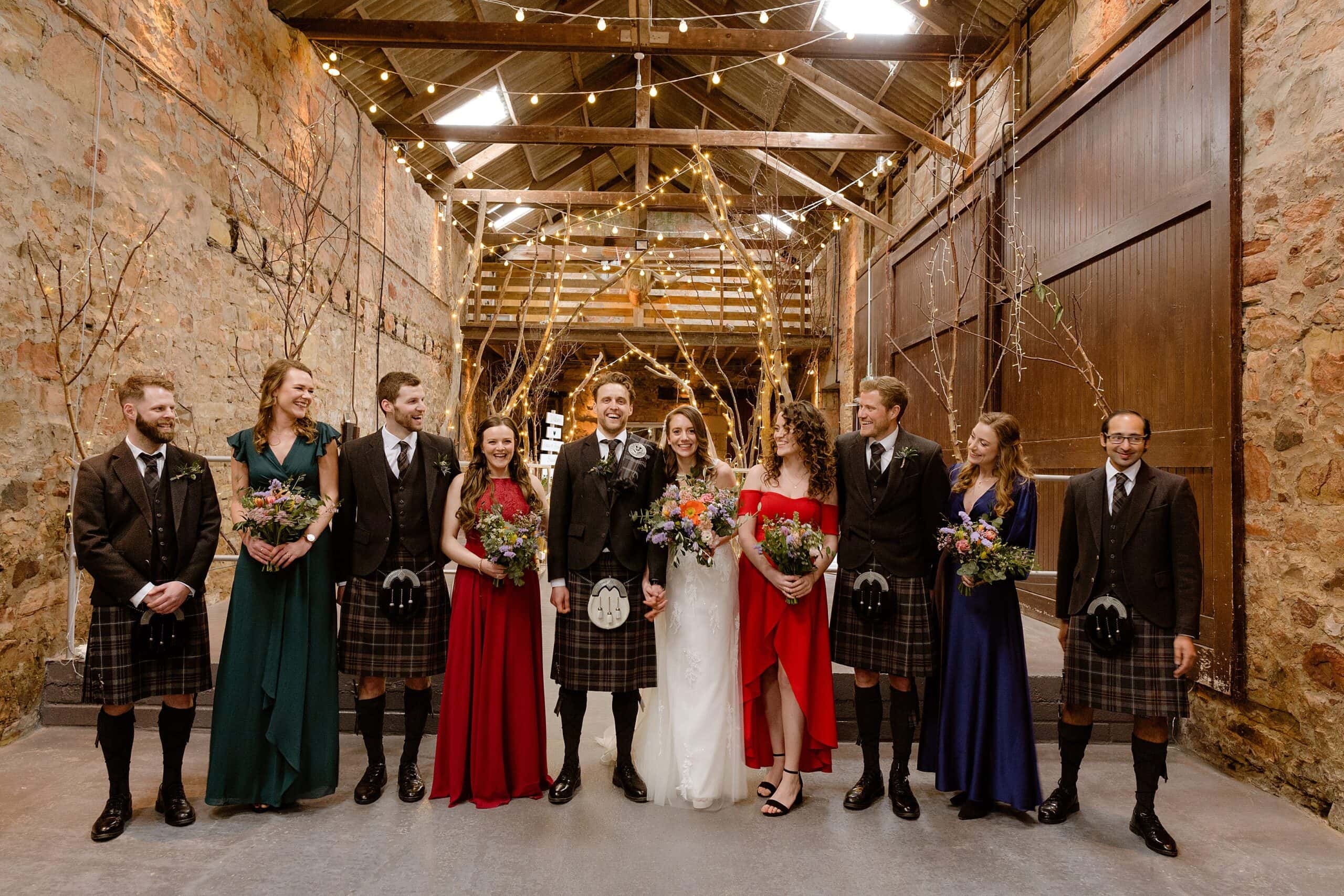 interior inside view of a farm wedding venue showing the bride groom bridesmaids and groomsmen standing in front of an archway of branches and festoon lights photographed by st andrews wedding photographer