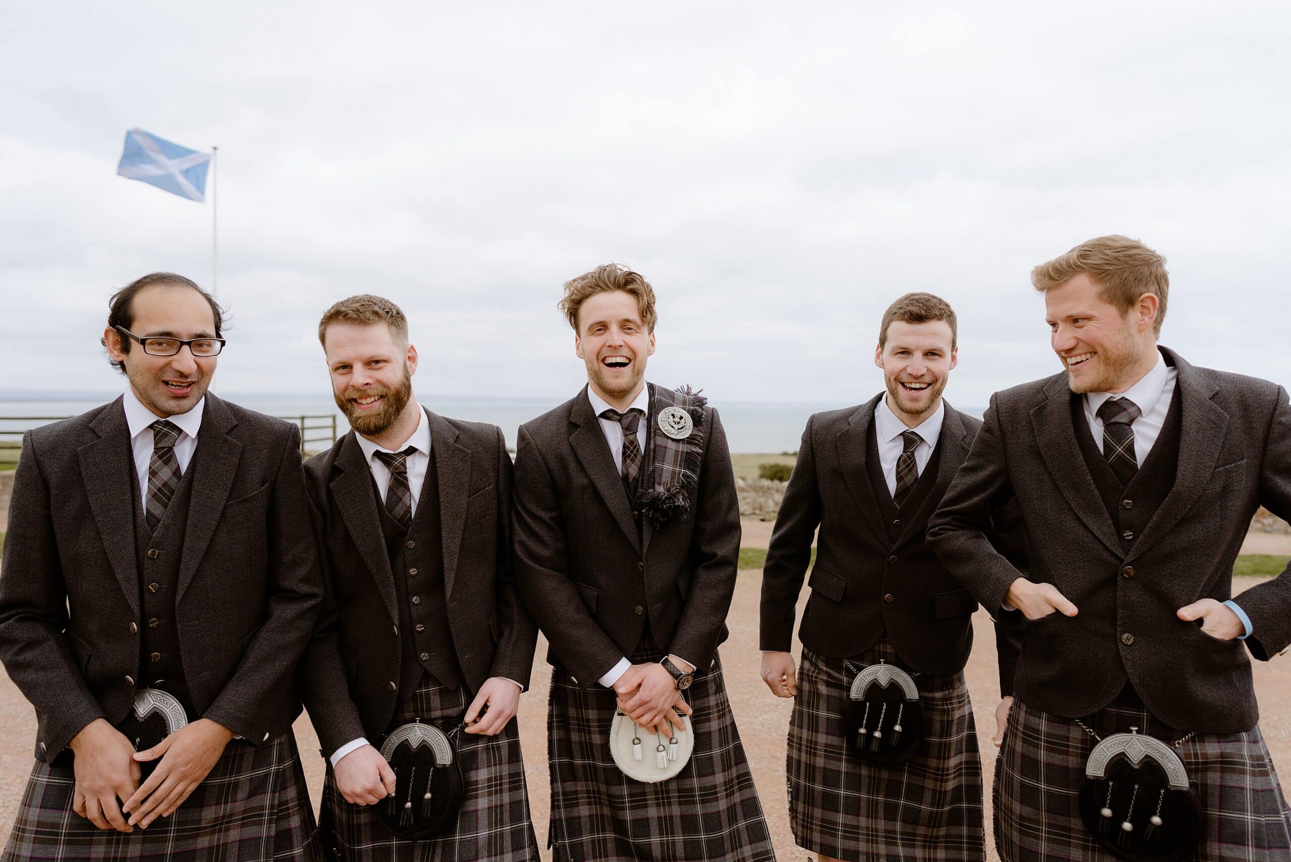 the groom and groomsmen standing outside a farm wedding venue in scotland with the sea and a scottish saltire flag in the background photographed by st andrews wedding photographer