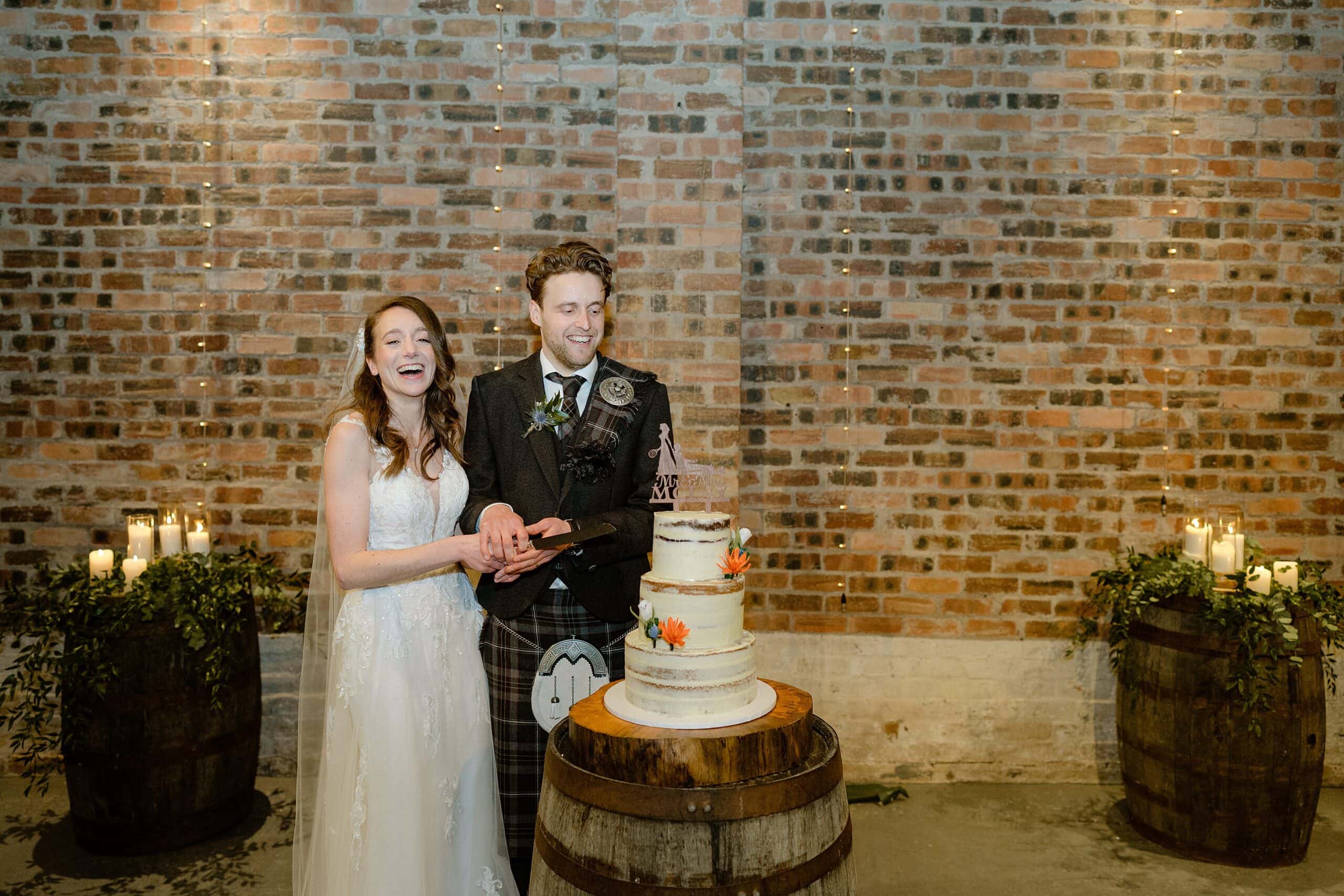 st andrews wedding photographer photographs the bride and groom smiling and cutting the wedding cake which sits on an oak barrel in front of a red brick wall and fairy lights