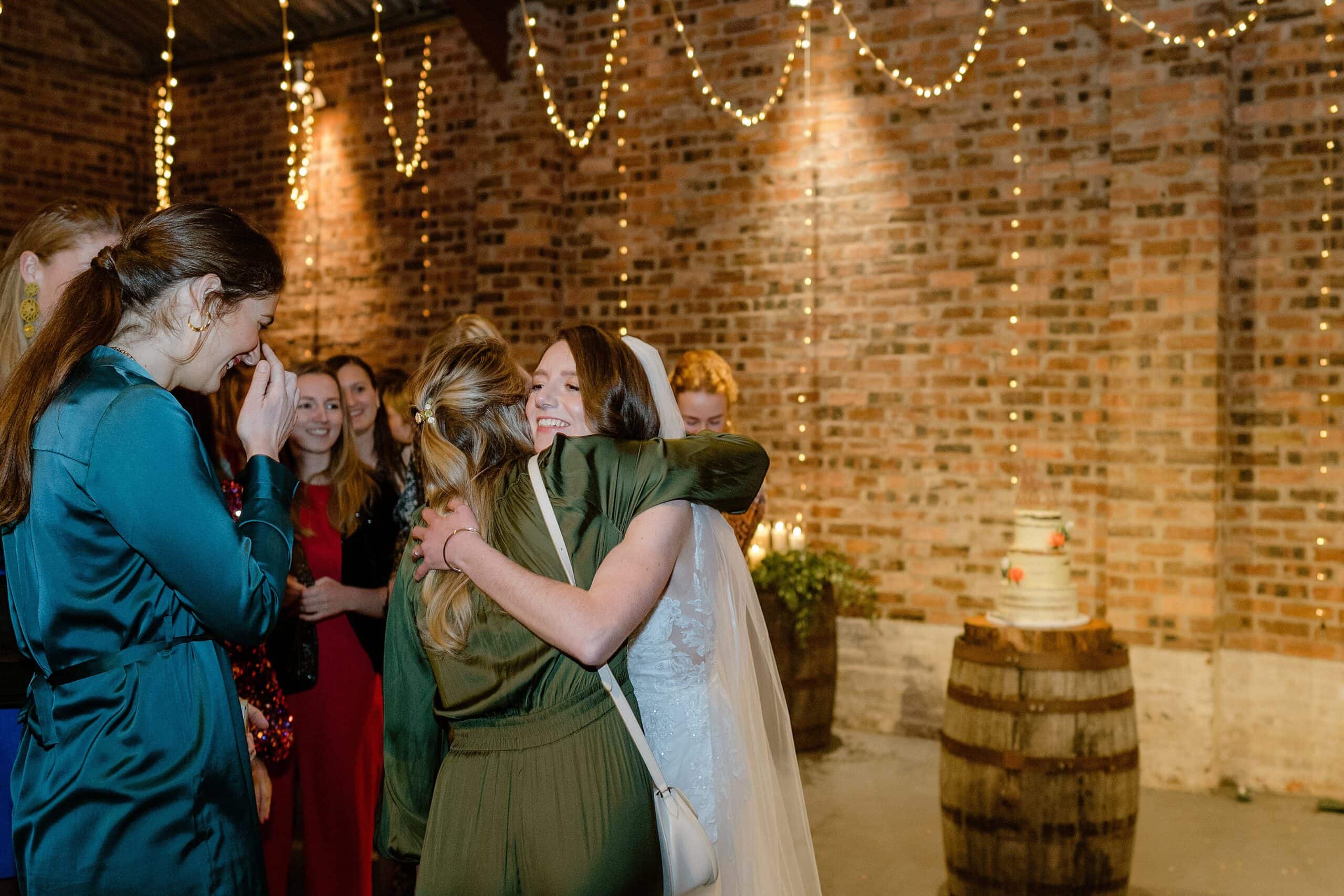 st andrews wedding photographer captures inside interior view of a barn wedding venue in scotland showing the bride hugging a guest following her wedding ceremony with the wedding cake sitting on an oak barrel and festoon lights behind