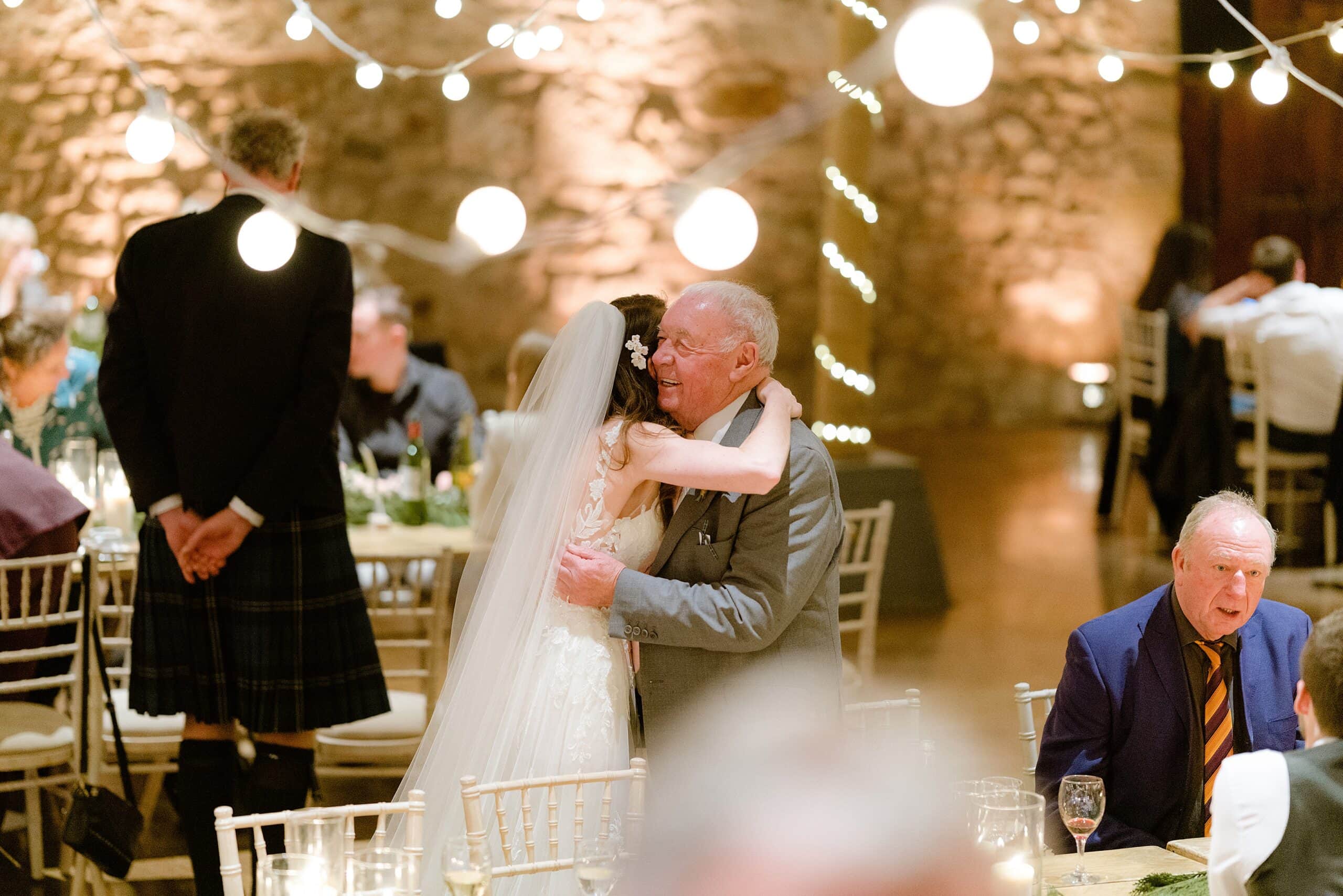 st andrews wedding photographer captures inside interior view a unique barn wedding venue in scotland showing the bride hugging a guest following her wedding ceremony with festoon lights above and in the background