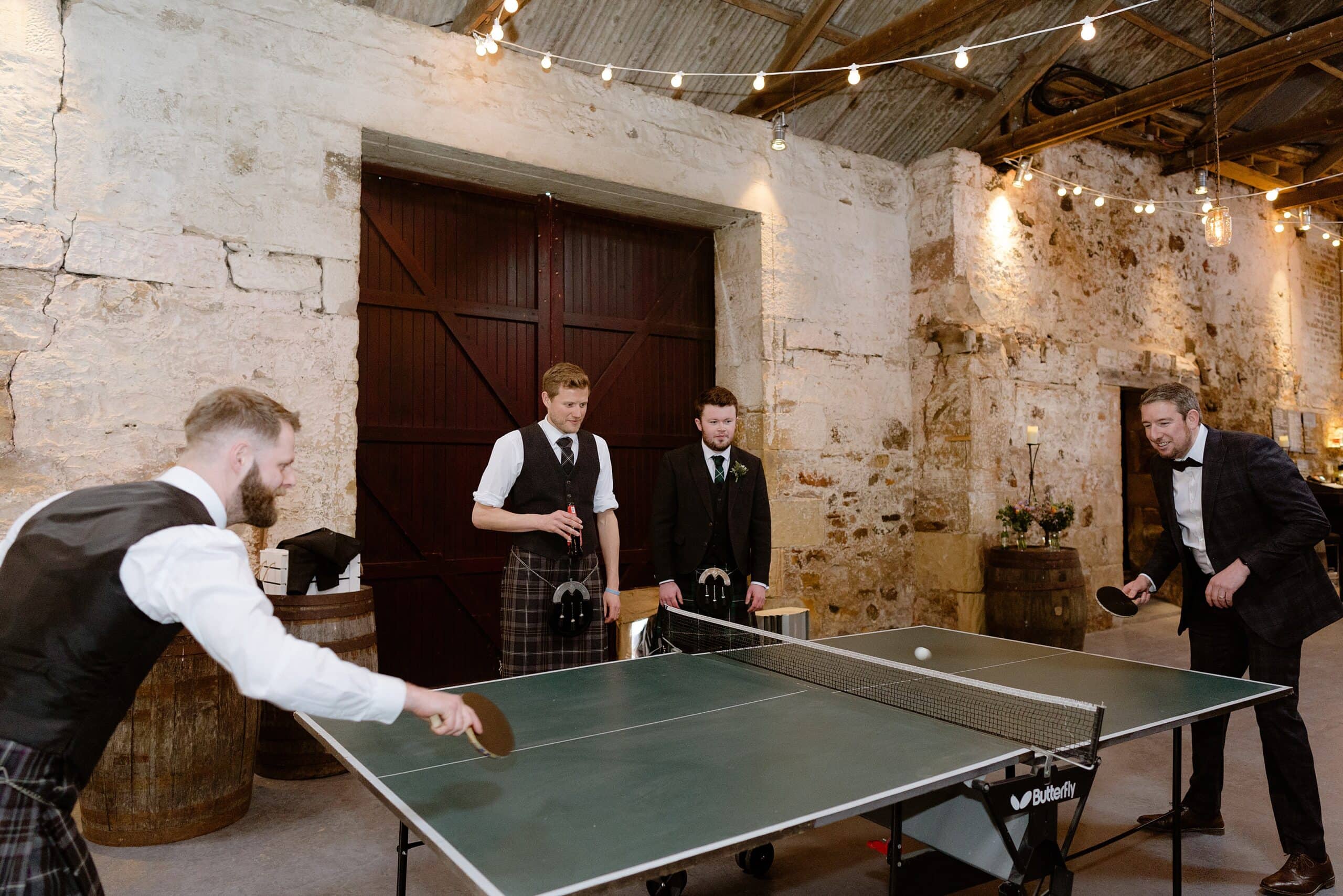 interior inside view of a scottish farm wedding venue showing guests playing table tennis with brick walls a barn door and oak barrels in the background and festoon lights above photographed by st andrews wedding photographer