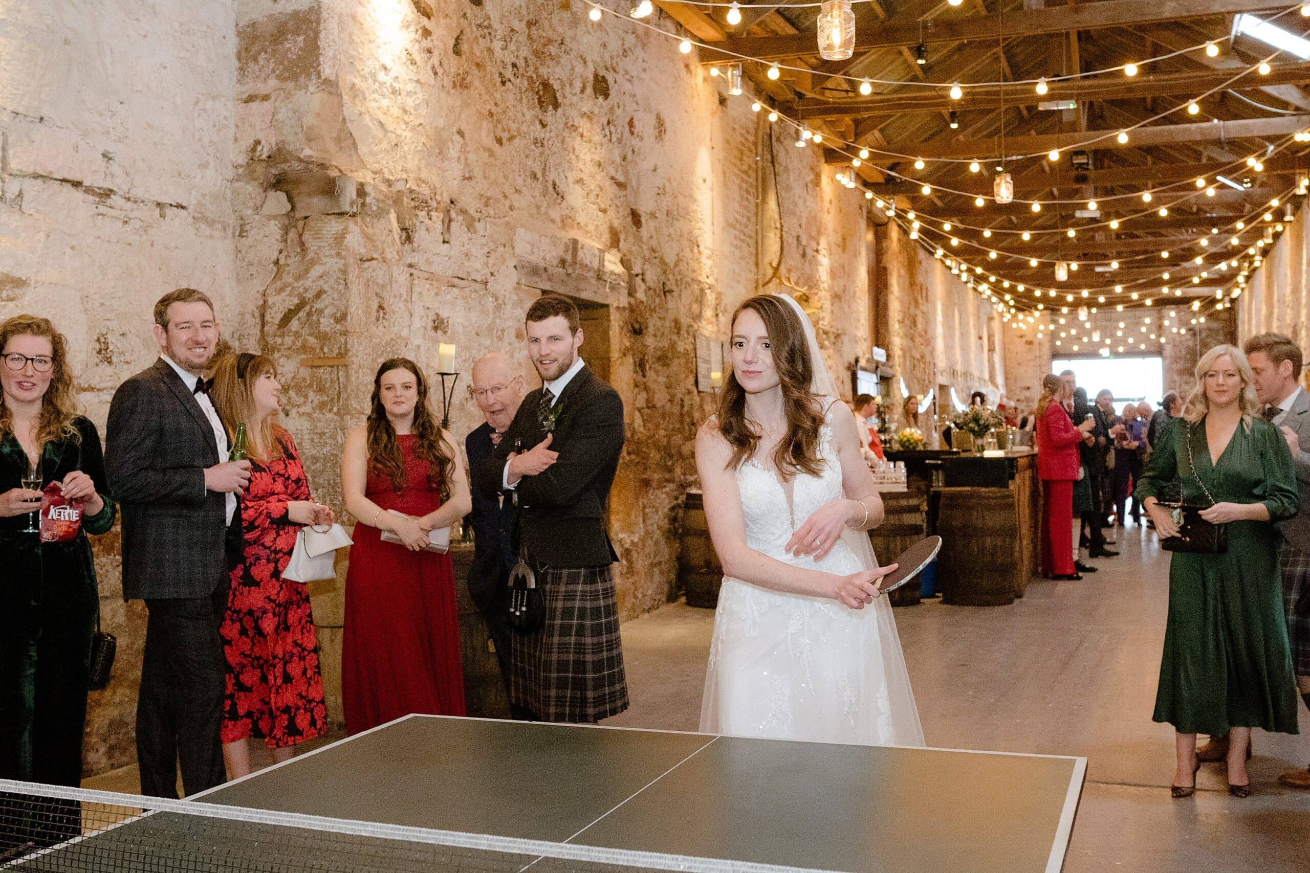 interior inside view of a scottish barn wedding venue showing the bride playing table tennis with festoon lights above and a bar in the background photographed by st andrews wedding photographer