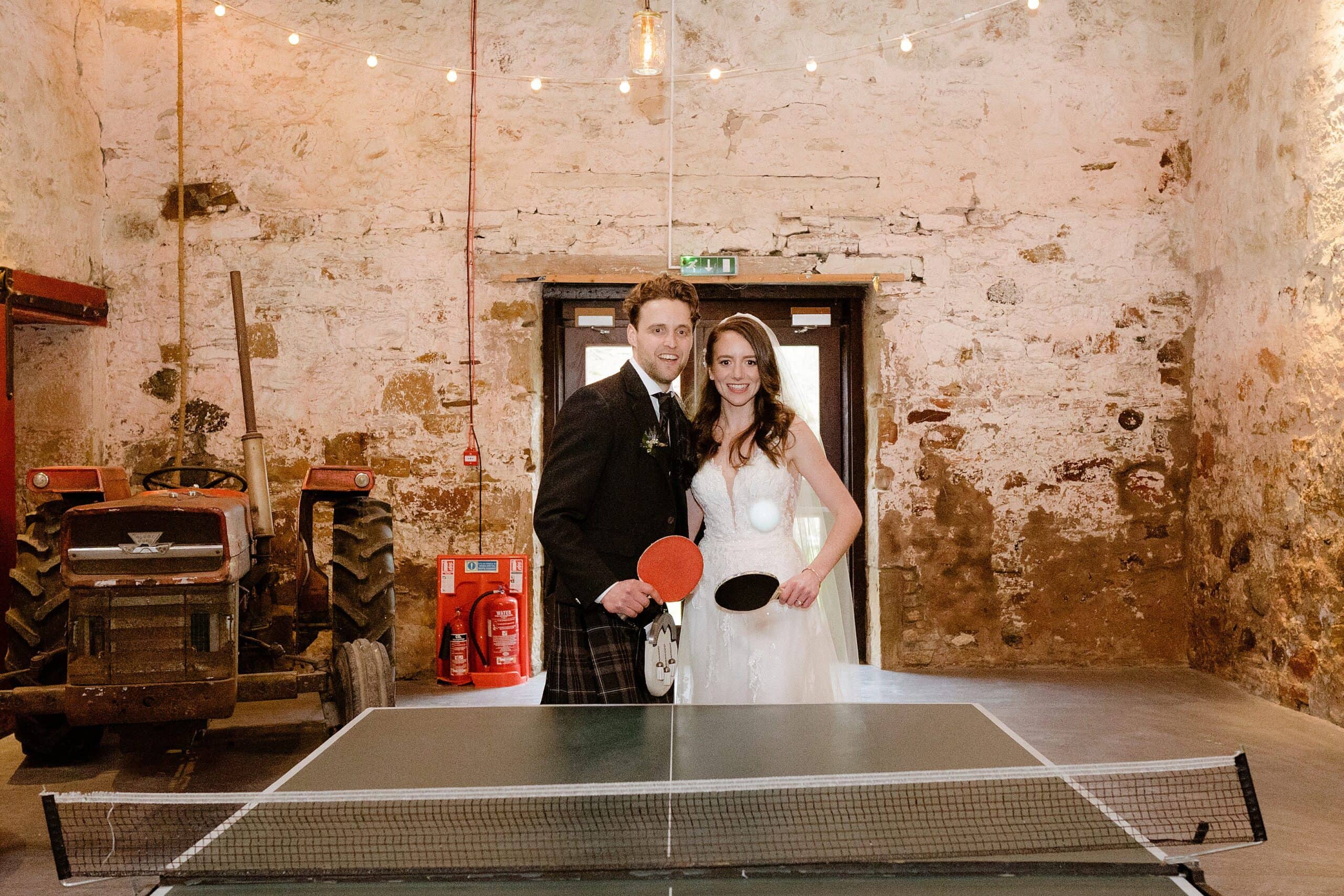 interior inside view of a unique barn wedding venue showing the bride and groom playing table tennis with festoon lights and a tractor in the background photographed by st andrews wedding photographer