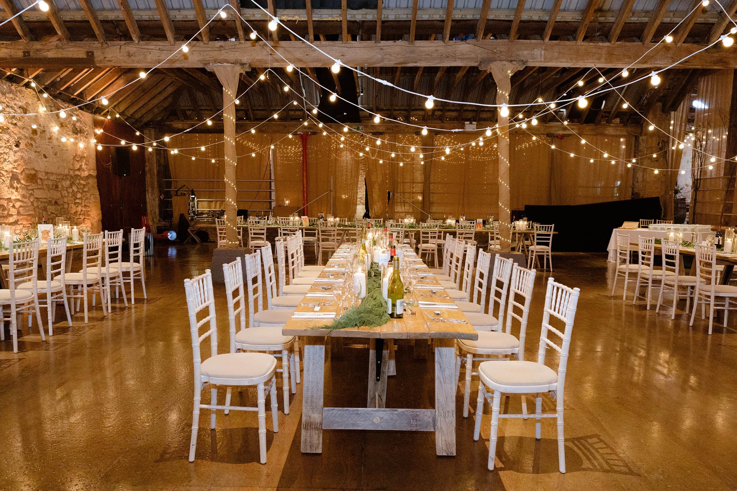 interior inside view of a scottish farm wedding venue with tables and chairs set for dinner with festoon lights and wooden beams above photographed by st andrews wedding photographer