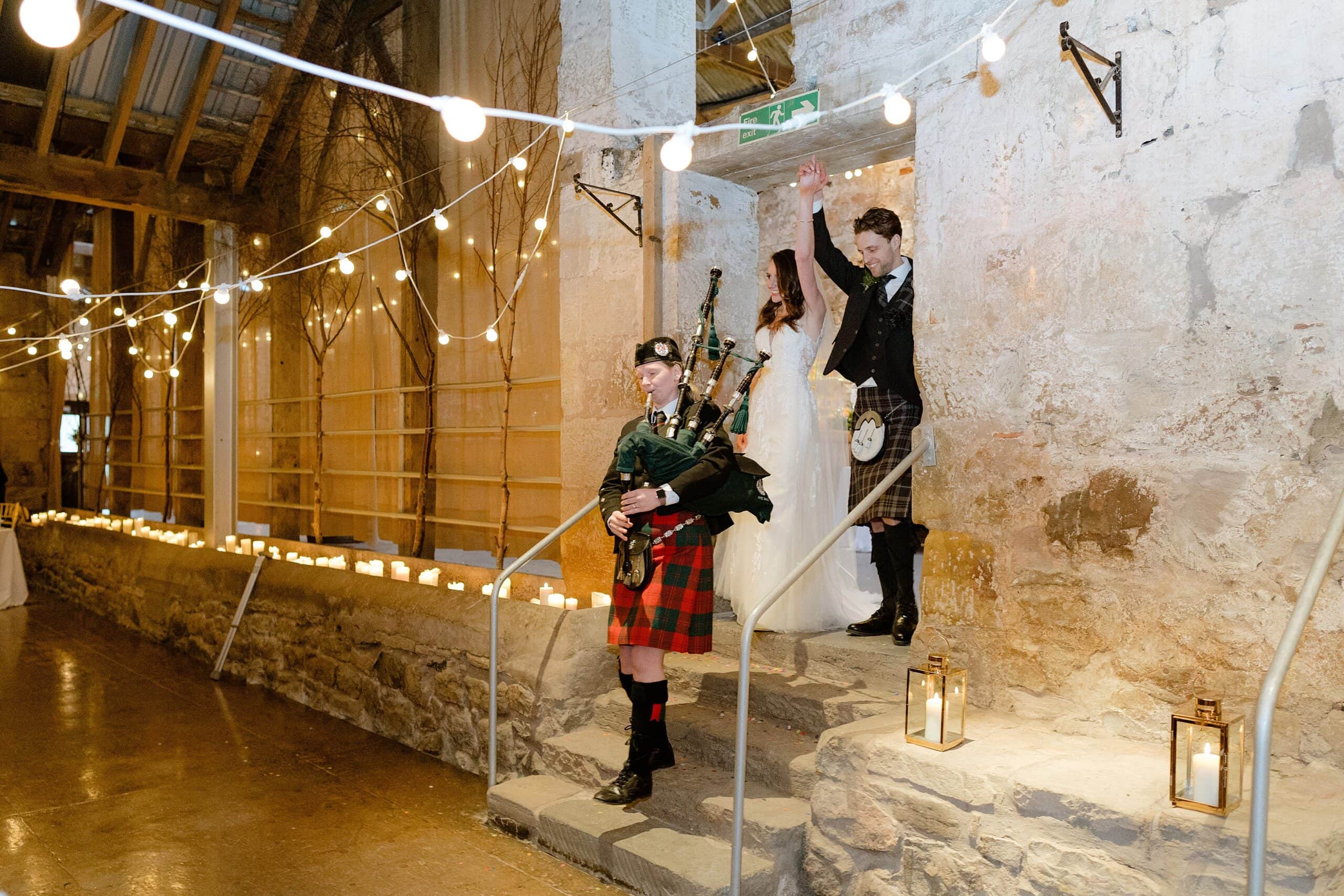 the bride and groom being piped in to dinner by a piper playing bagpipes with festoon lights and candles in the background captured by st andrews wedding photographer