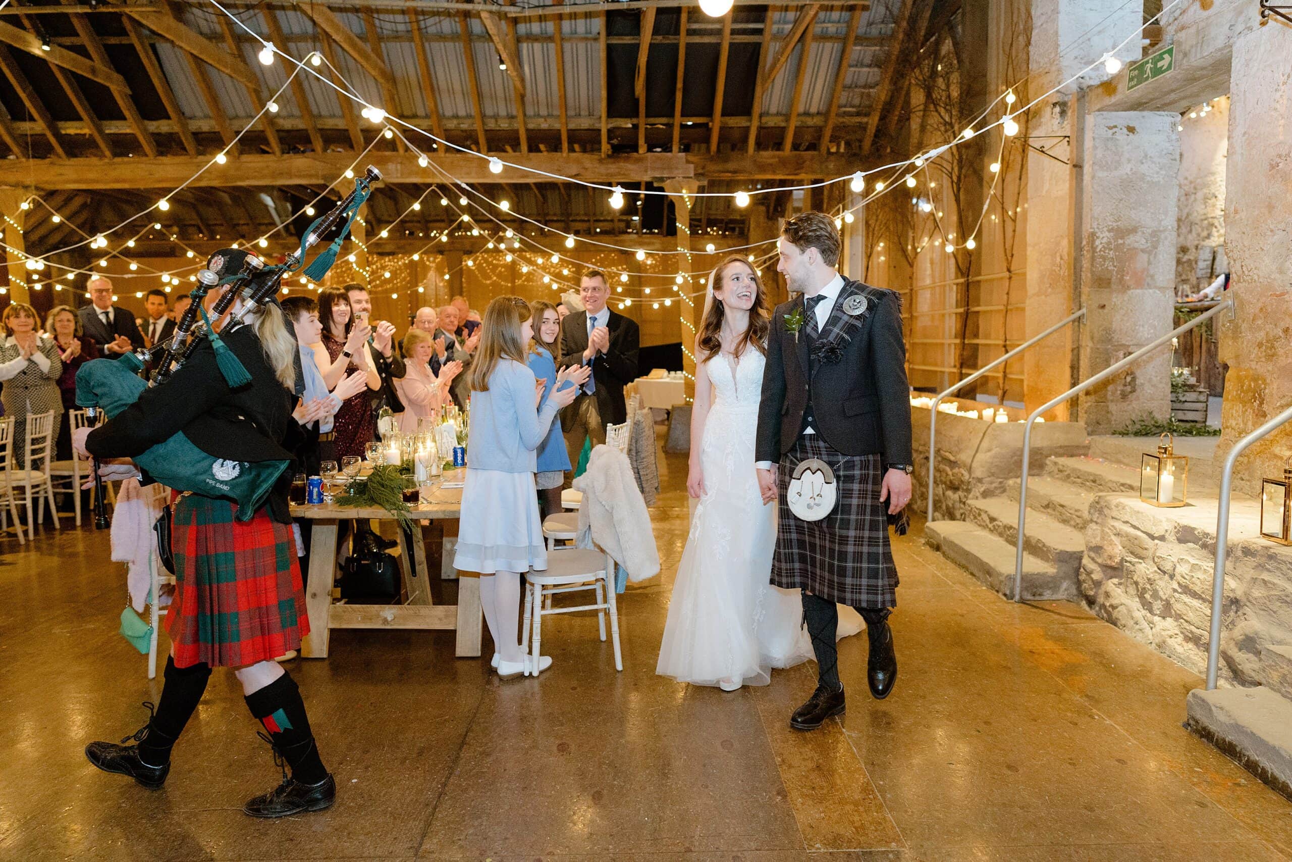 the bride and groom being piped in to dinner by a piper playing bagpipes with festoon lights and candles in the background captured by st andrews wedding photographer
