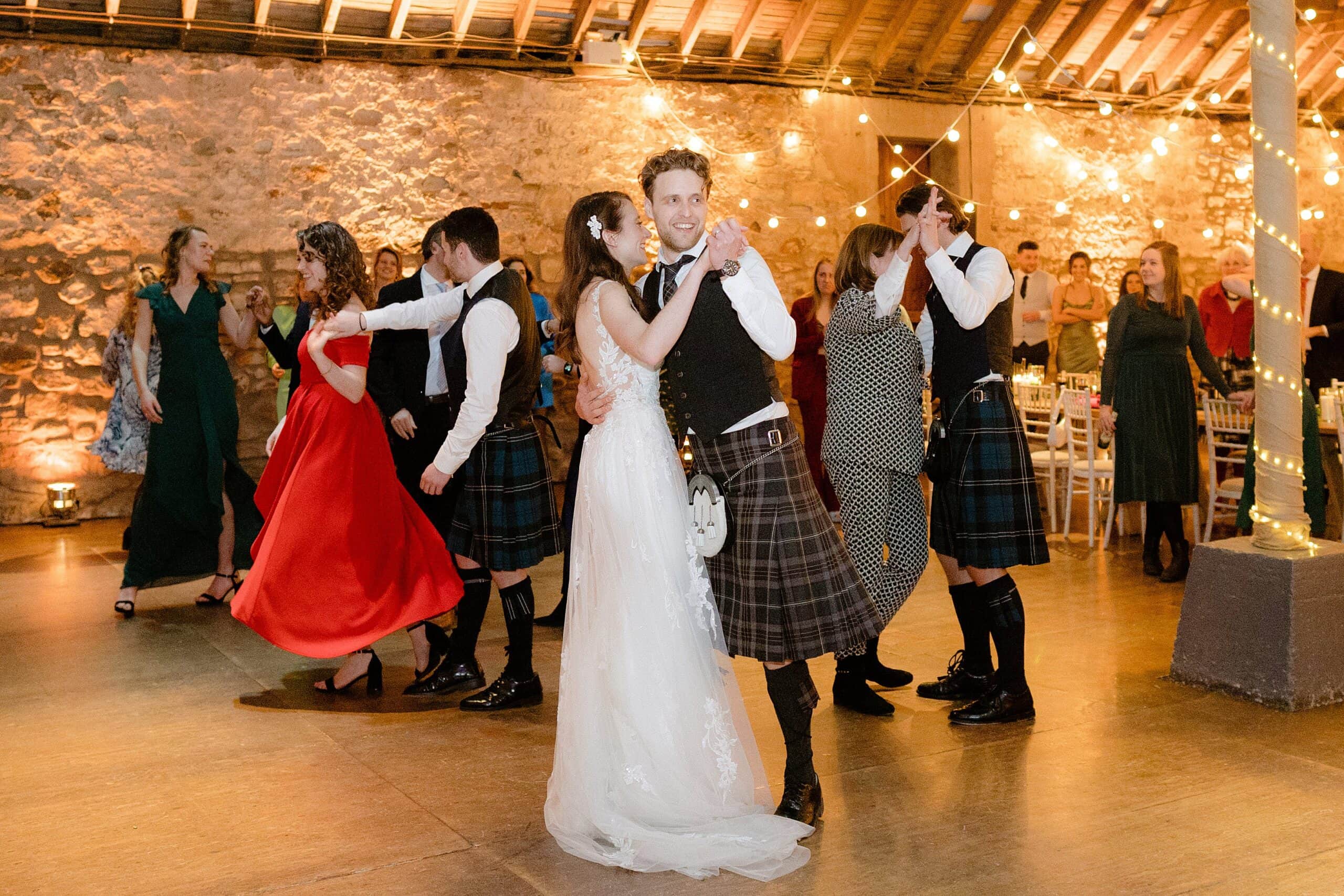 interior inside view of a barn wedding venue in scotland showing the bride groom and guests dancing underneath wooden beams and festoon lights captured by st andrews wedding photographer