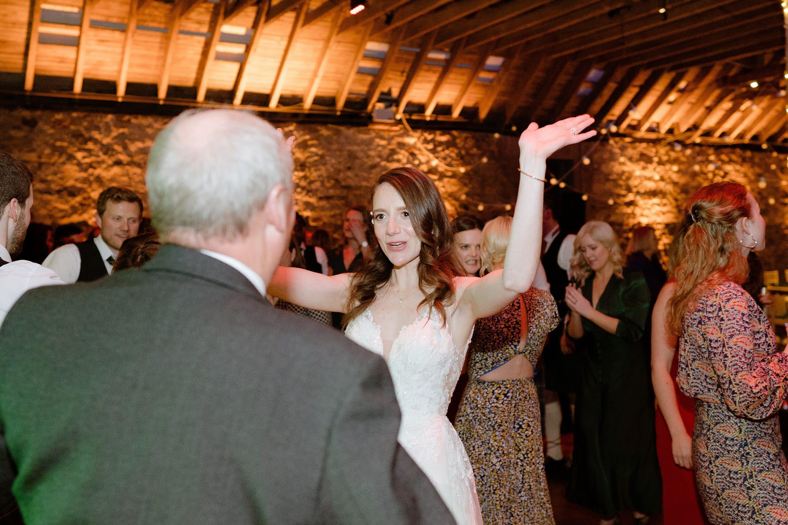 st andrews wedding photographer captures the bride dancing with a guest underneath wooden beams with festoon lights in the background at a barn wedding venue in scotland
