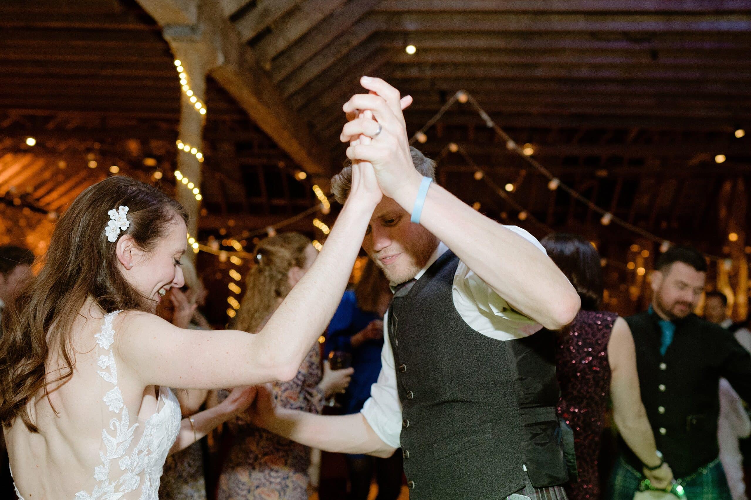 st andrews wedding photographer captures the bride dancing with a guest underneath wooden beams with festoon lights in the background at a farm wedding venue in scotland