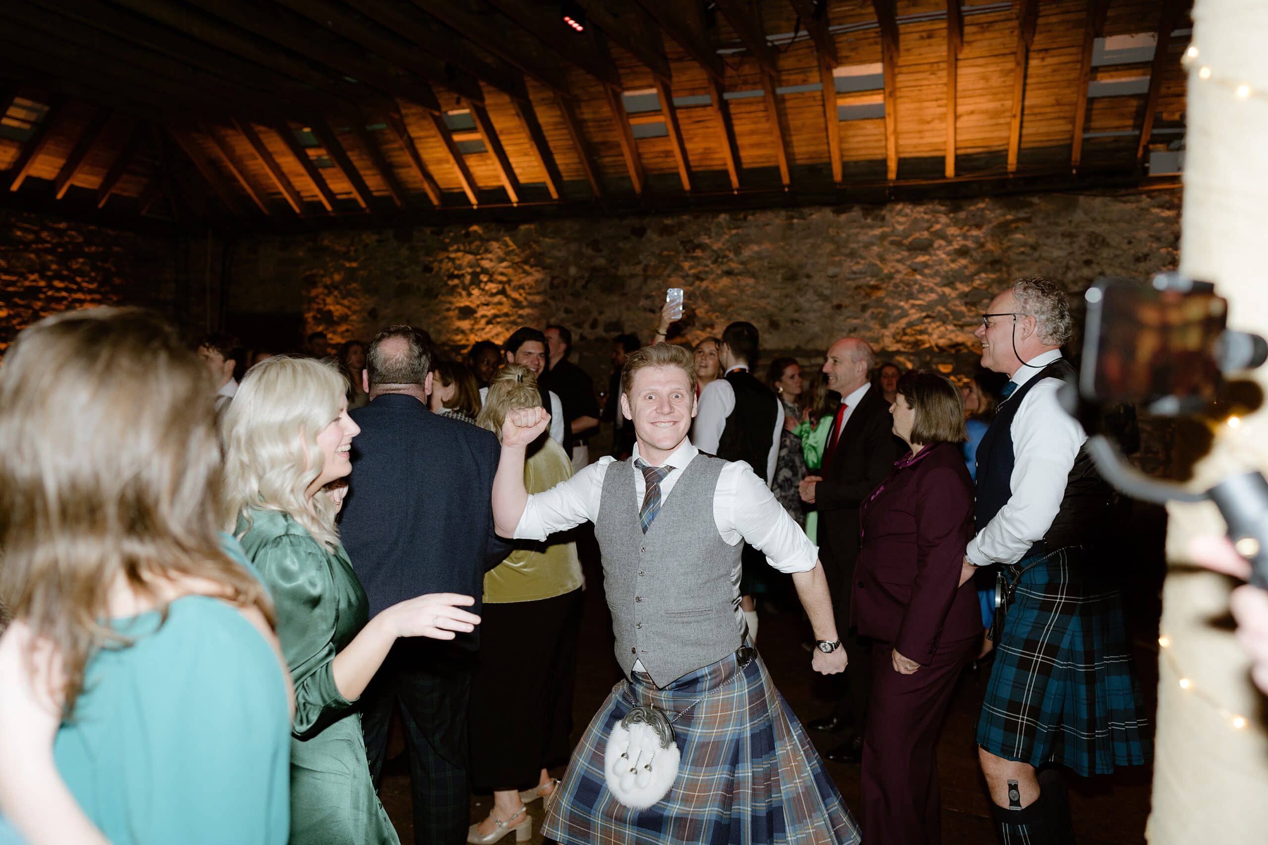 st andrews wedding photographer captures guests dancing underneath wooden beams and festoon lights a barn wedding venue in scotland